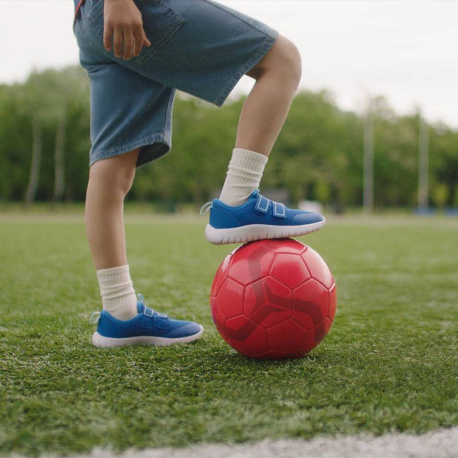 Gyerek rövidnadrágban és kék sportcipőben, egyik lábával egy piros focilabdán.