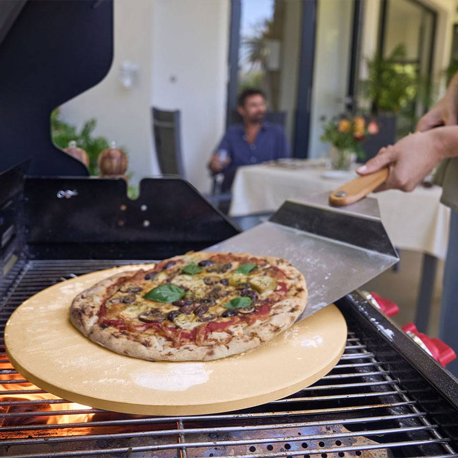 Pizza pizzakövön grillen, pizzalapáttal és egy férfival a háttérben.