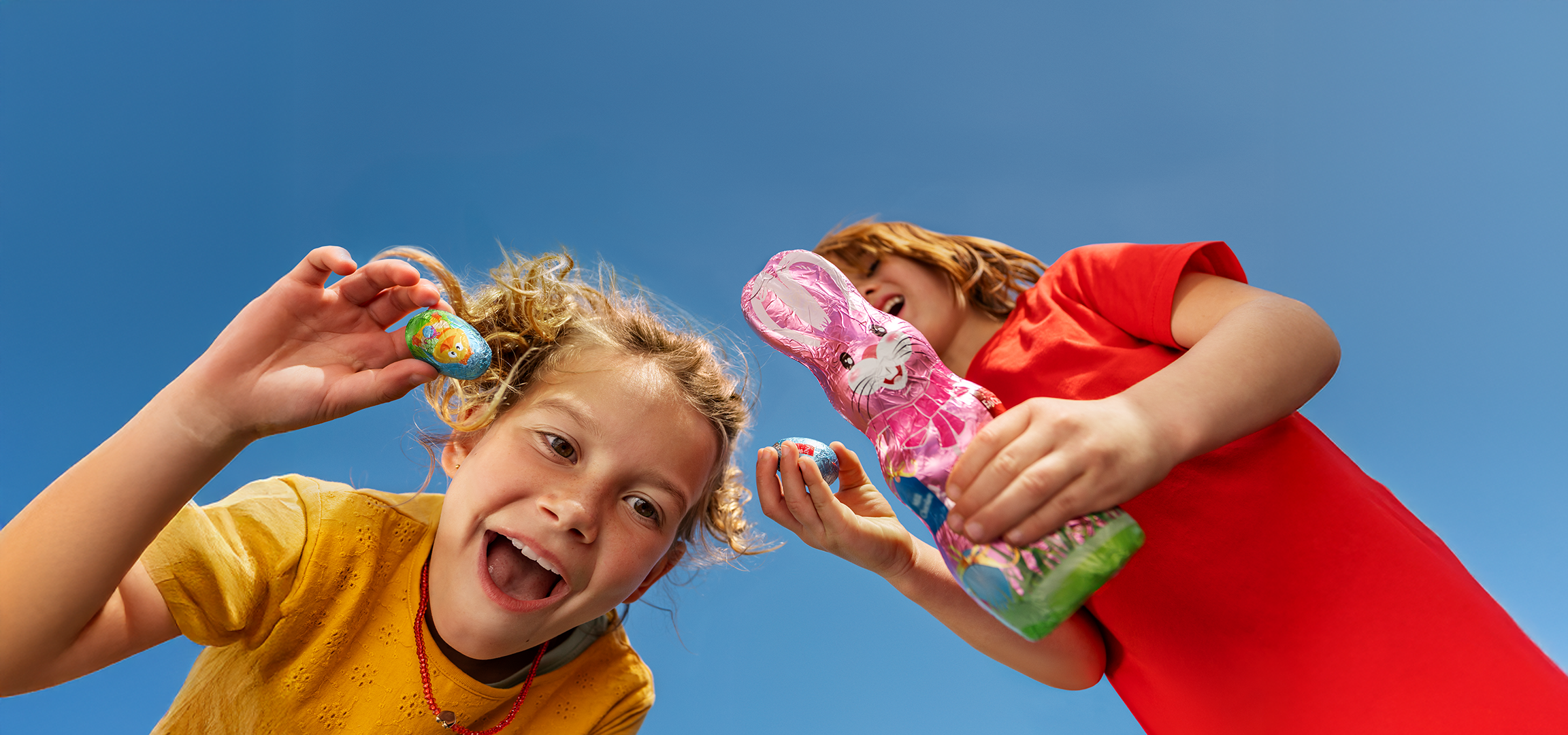 Gyerekek húsvéti csokitojásokkal és nyuszikkal a kék ég alatt.