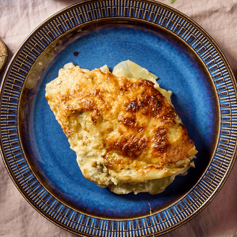 Sült rakott étel kék tányéron, felül aranybarnára pirult sajttal.