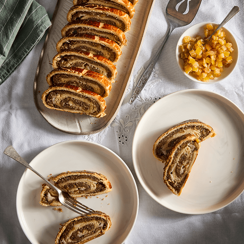 Szeletelt mákos bejgli tálalva, kandírozott gyümölcsökkel és villával.