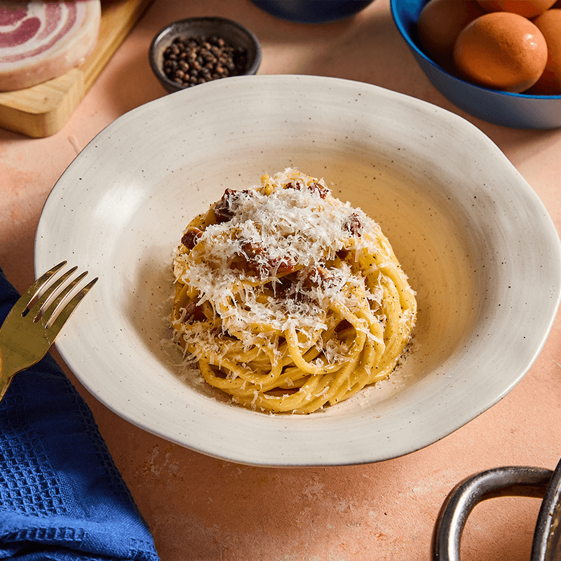 Spagetti carbonara szalonnával, parmezánnal és fekete borssal, tojásokkal a háttérben.