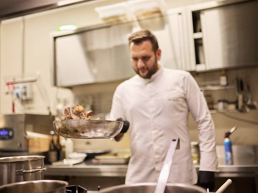 Fehér szakácskabátos séf ételt dobál egy serpenyőben.