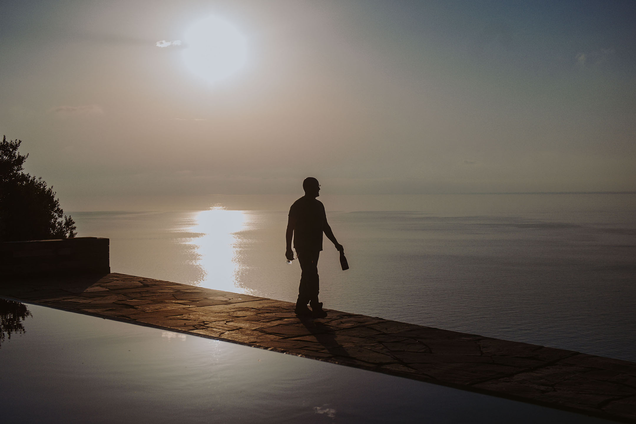 Egy személy sziluettje palackkal és pohárral egy végtelenített medence mellett naplementekor