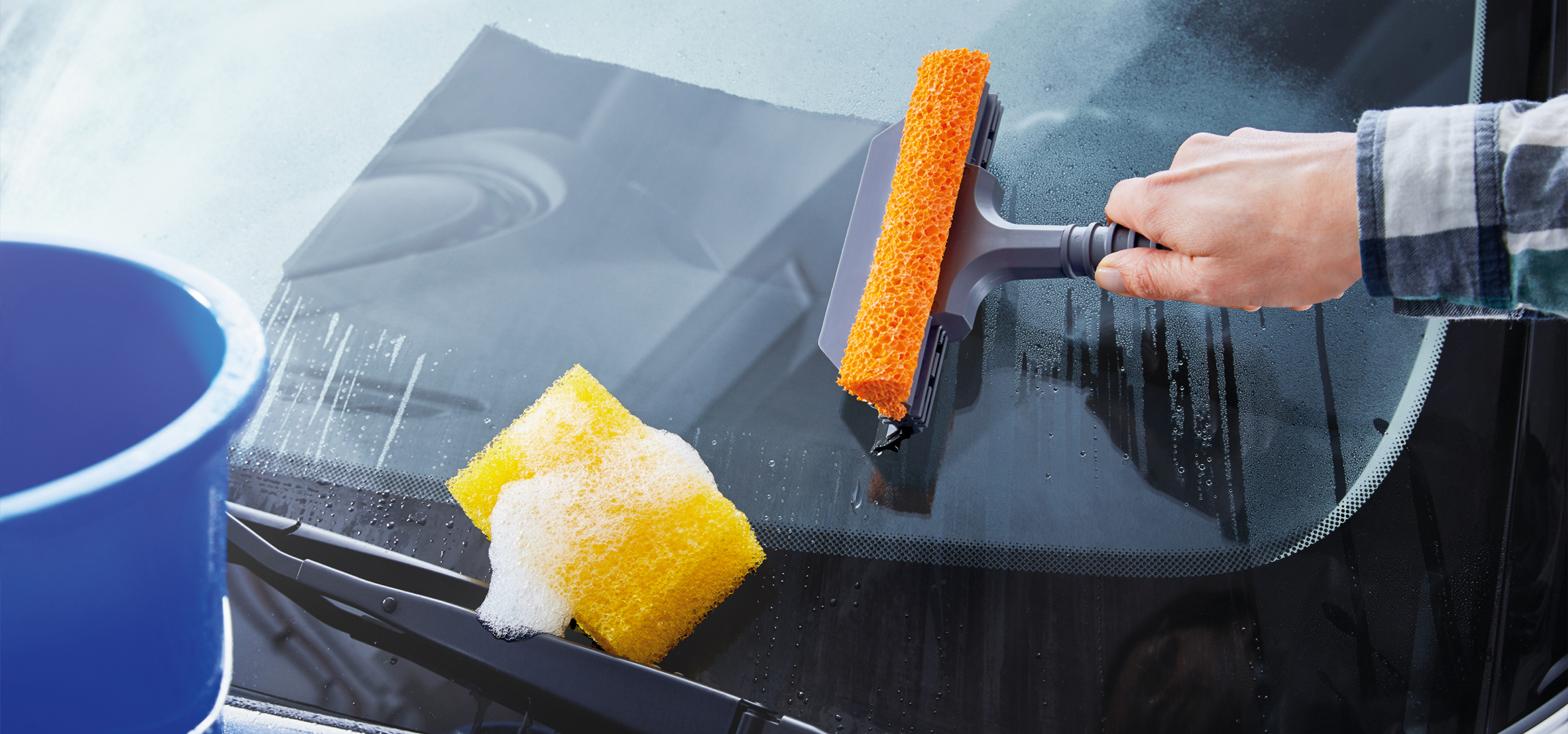 Kéz tisztít egy autó szélvédőjét egy narancssárga szivaccsal és egy sárga szivaccsal.