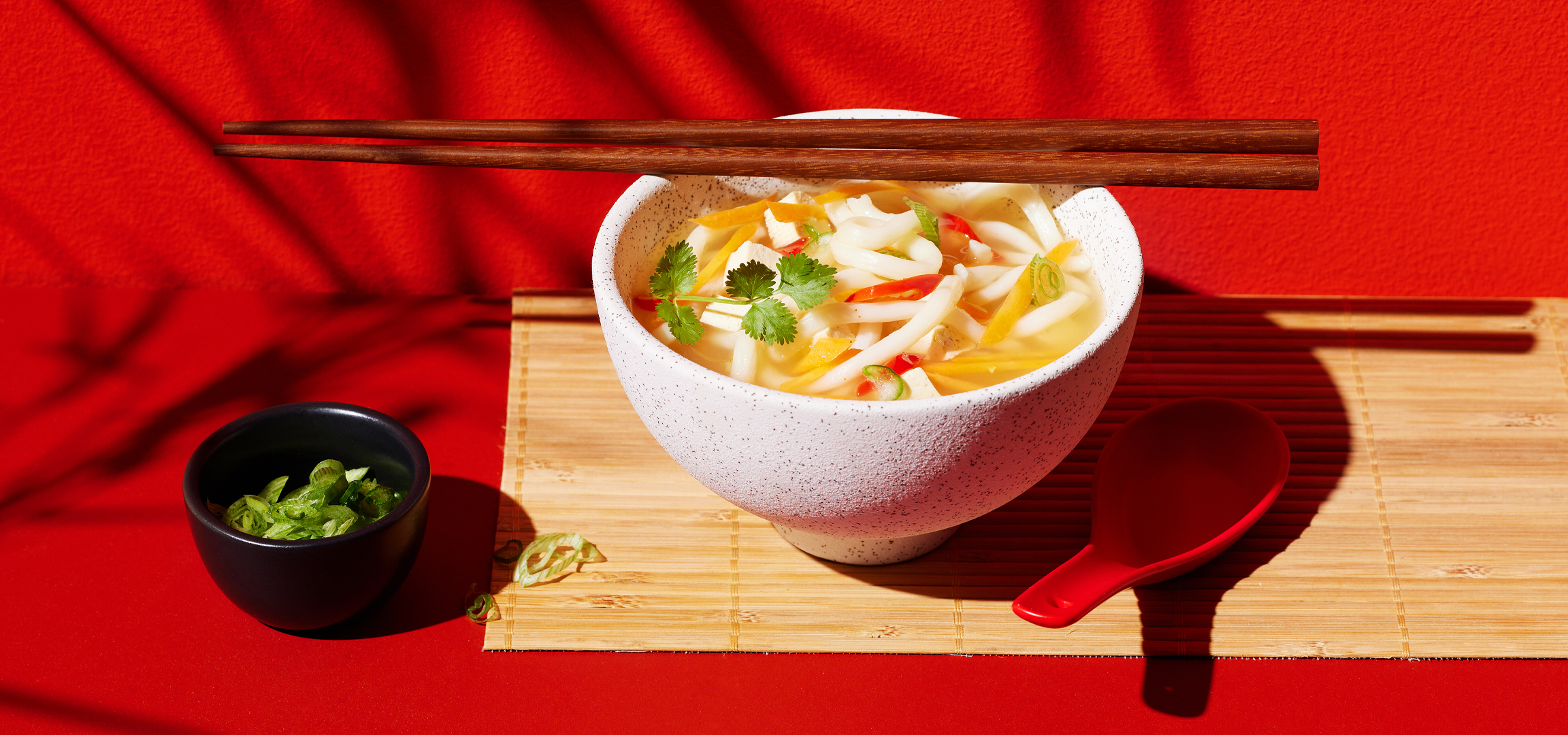 Ázsiai tésztaétel tofuval és zöldségekkel, pálmalevél árnyékokkal egy piros asztalon.