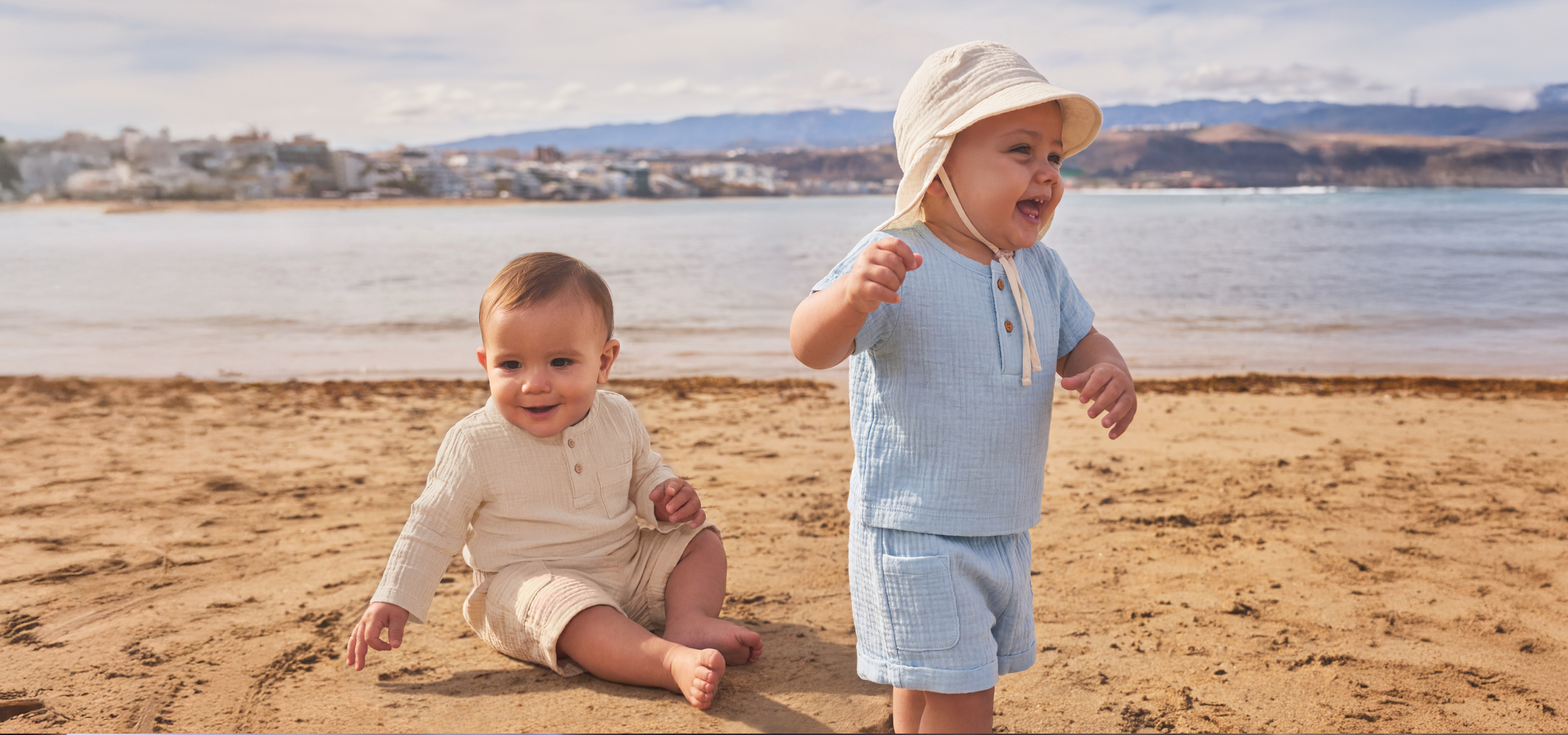 Két kisgyermek játszik a homokos tengerparton, a háttérben a tenger és egy város látható.