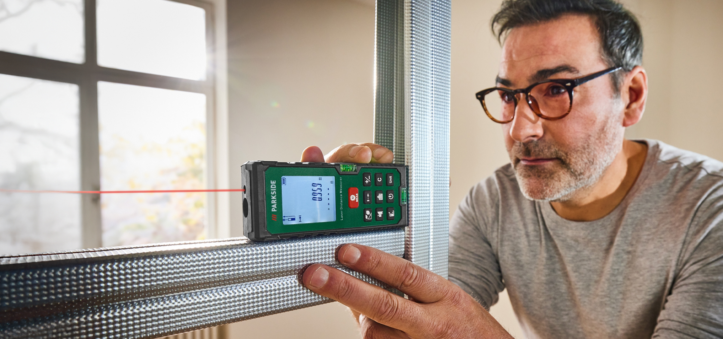 Férfi lézeres távolságmérővel mér egy fémprofilt, háttérben ablak.