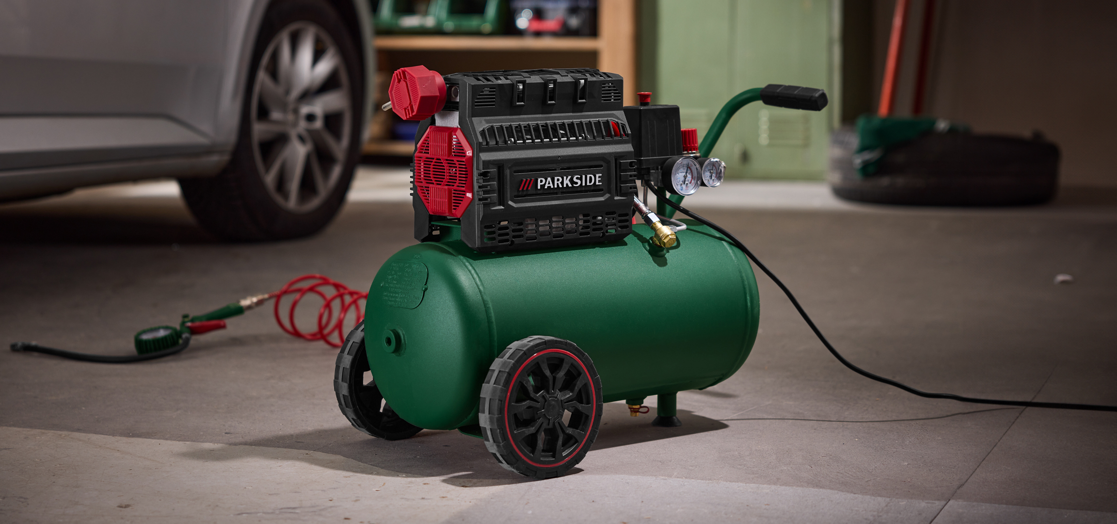 Zöld kompresszor piros tömlővel és mérőórákkal egy ezüst autó mellett egy garázsban.