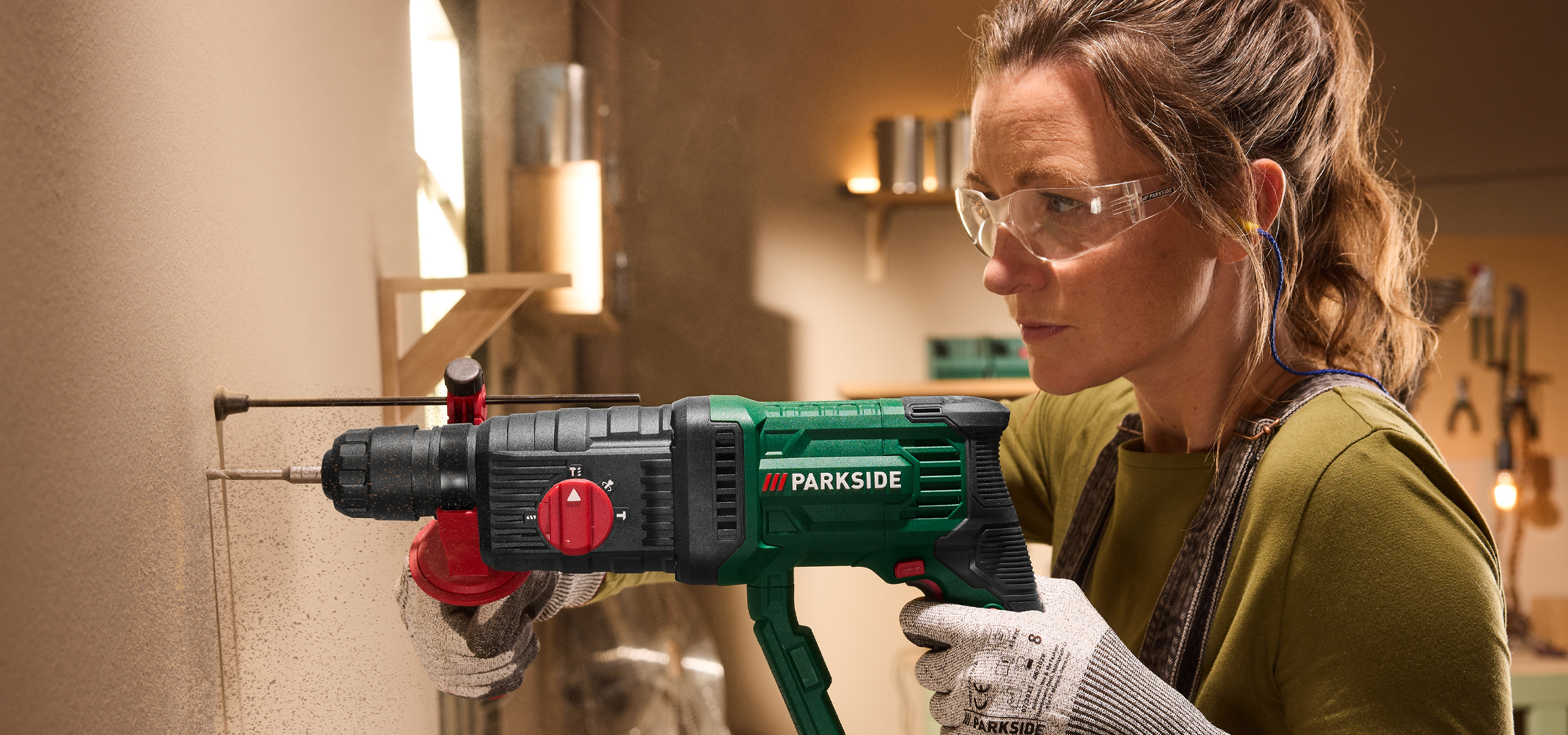 Nő fúrógéppel fúr egy falba, védőszemüveget és fülvédőt visel.