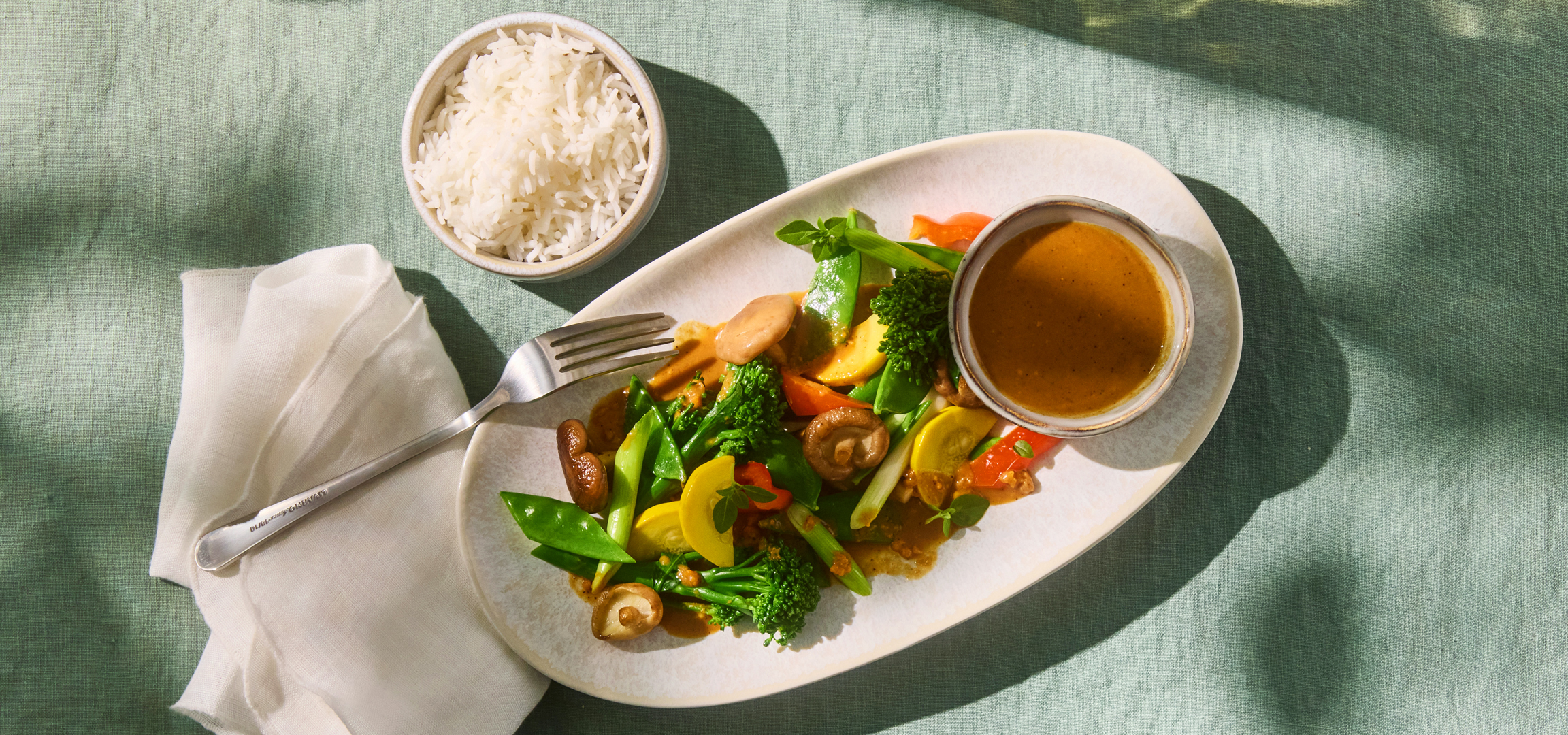 Zöldséges stir-fry mogyorószószban, rizzsel és villával, zöld terítőn.