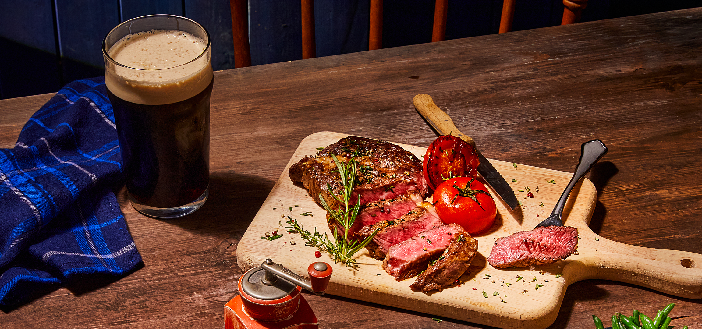 Sült steak paradicsommal és zöldbabbal, sörrel és kék kockás szalvétával egy fa asztalon.