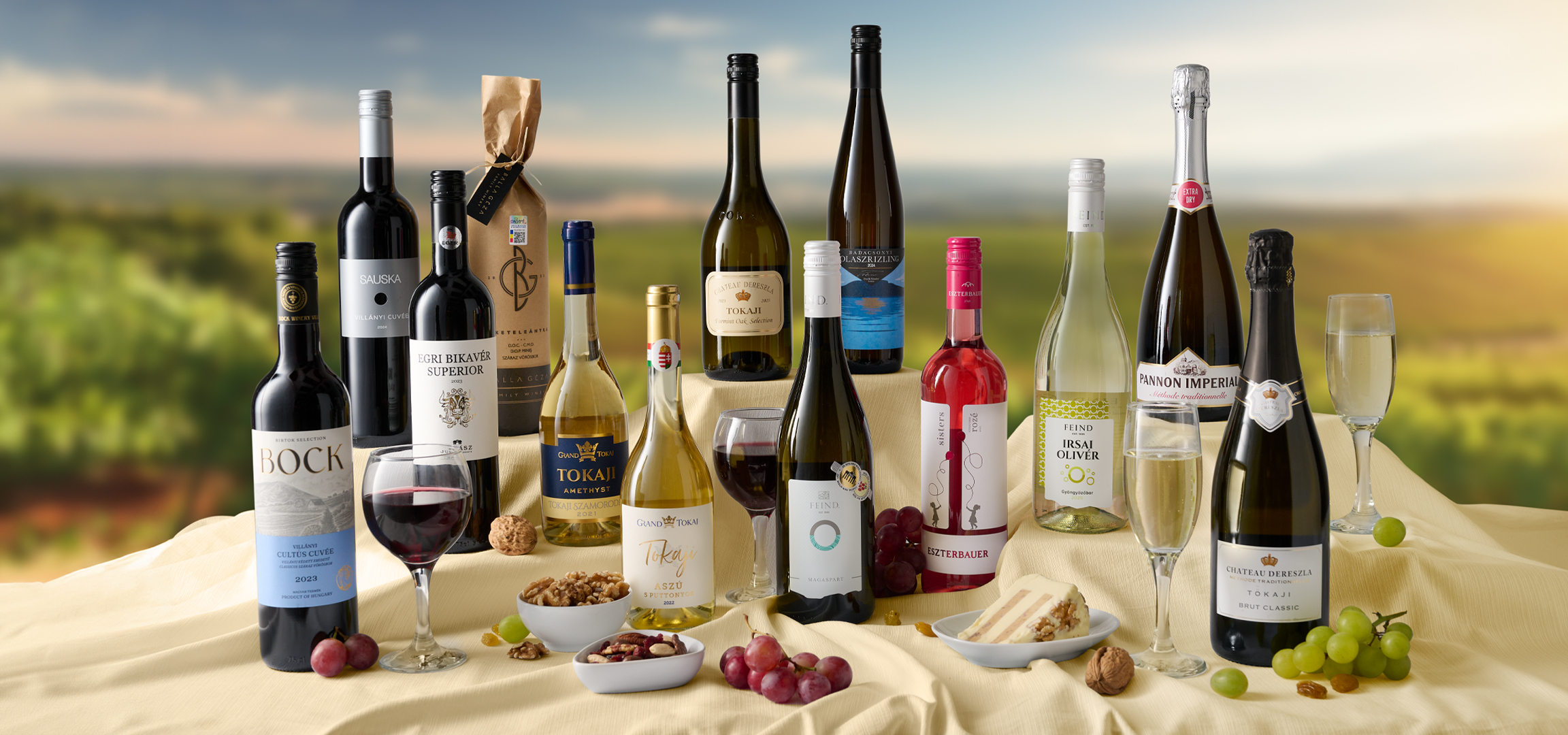 Különböző borok és pezsgők egy szőlőültetvény előtt, dióval, szőlővel és süteménnyel.