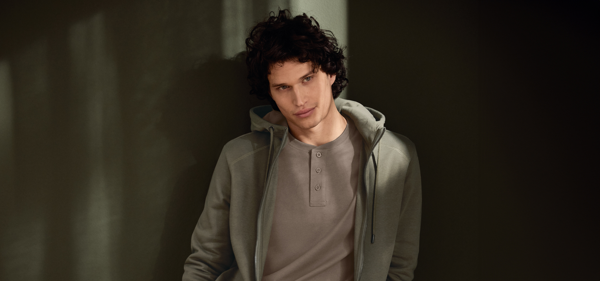 Fiatal férfi kapucnis pulóverben és Henley pólóban, sötét háttér előtt.