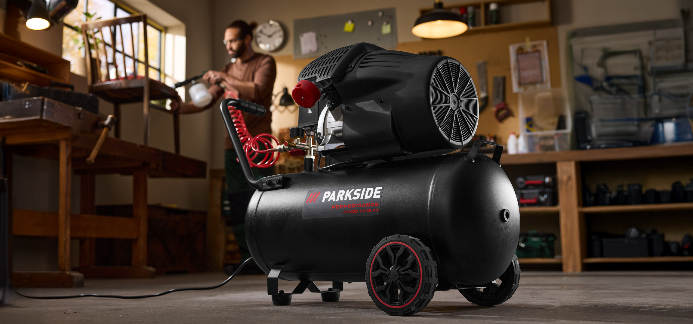 Fekete kompresszor egy műhelyben, háttérben egy férfi dolgozik egy széken.