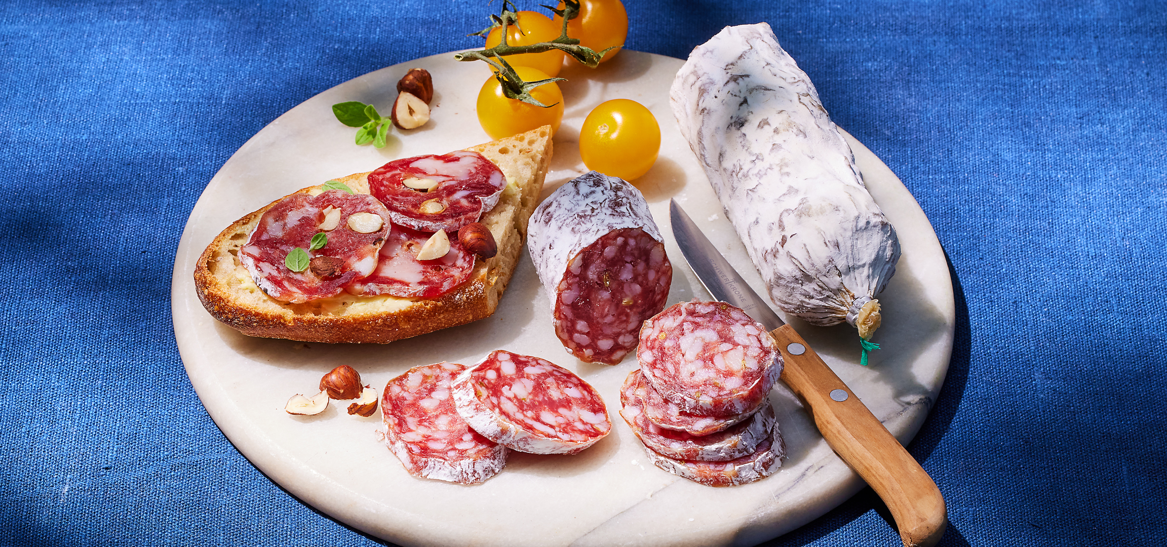 Szalámi bruschetta, egész szalámi, sárga koktélparadicsom és mogyoró márványtálon.