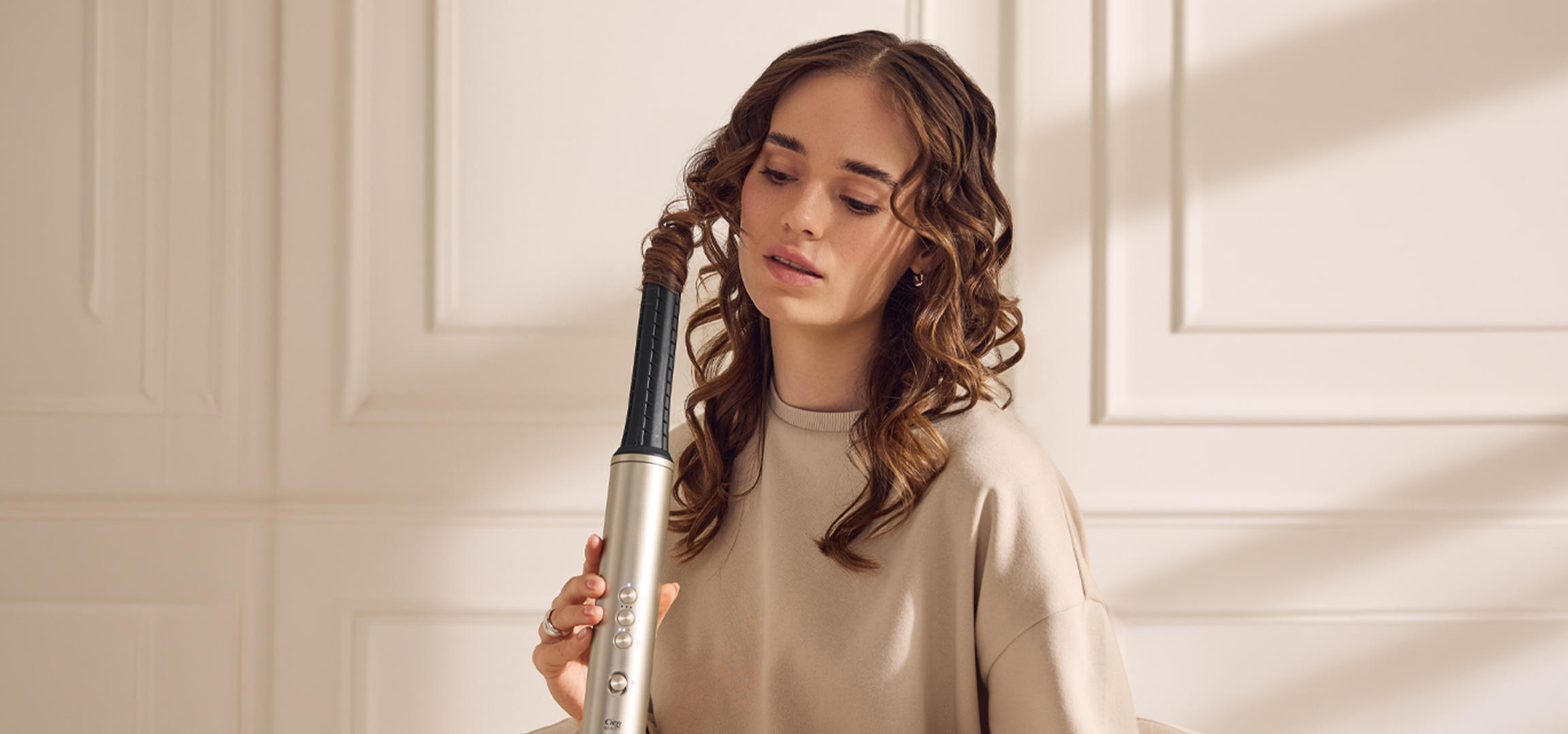 Fiatal nő göndöríti a haját egy hajgöndörítővel, otthoni környezetben.