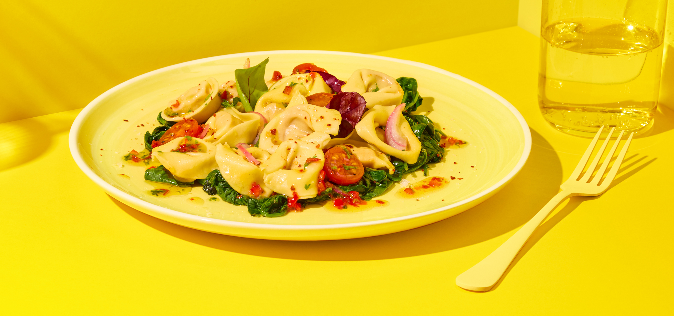 Tortellini spenóttal és paradicsommal, sárga tányéron, sárga háttér előtt.