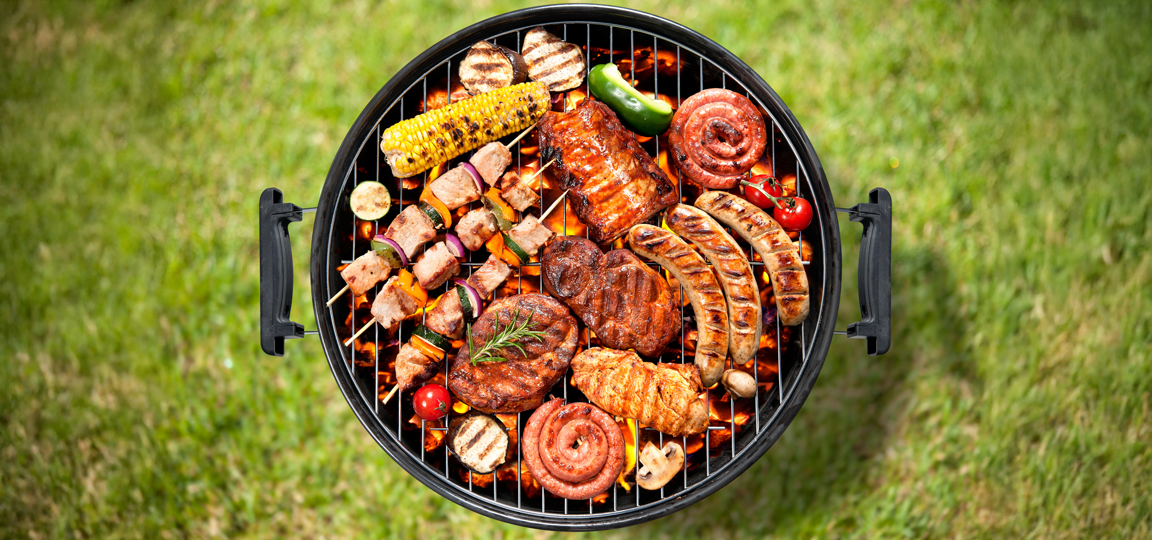 Grillezett húsok, kolbászok, kukorica és zöldségek egy kerti grillen, zöld fű háttérrel.