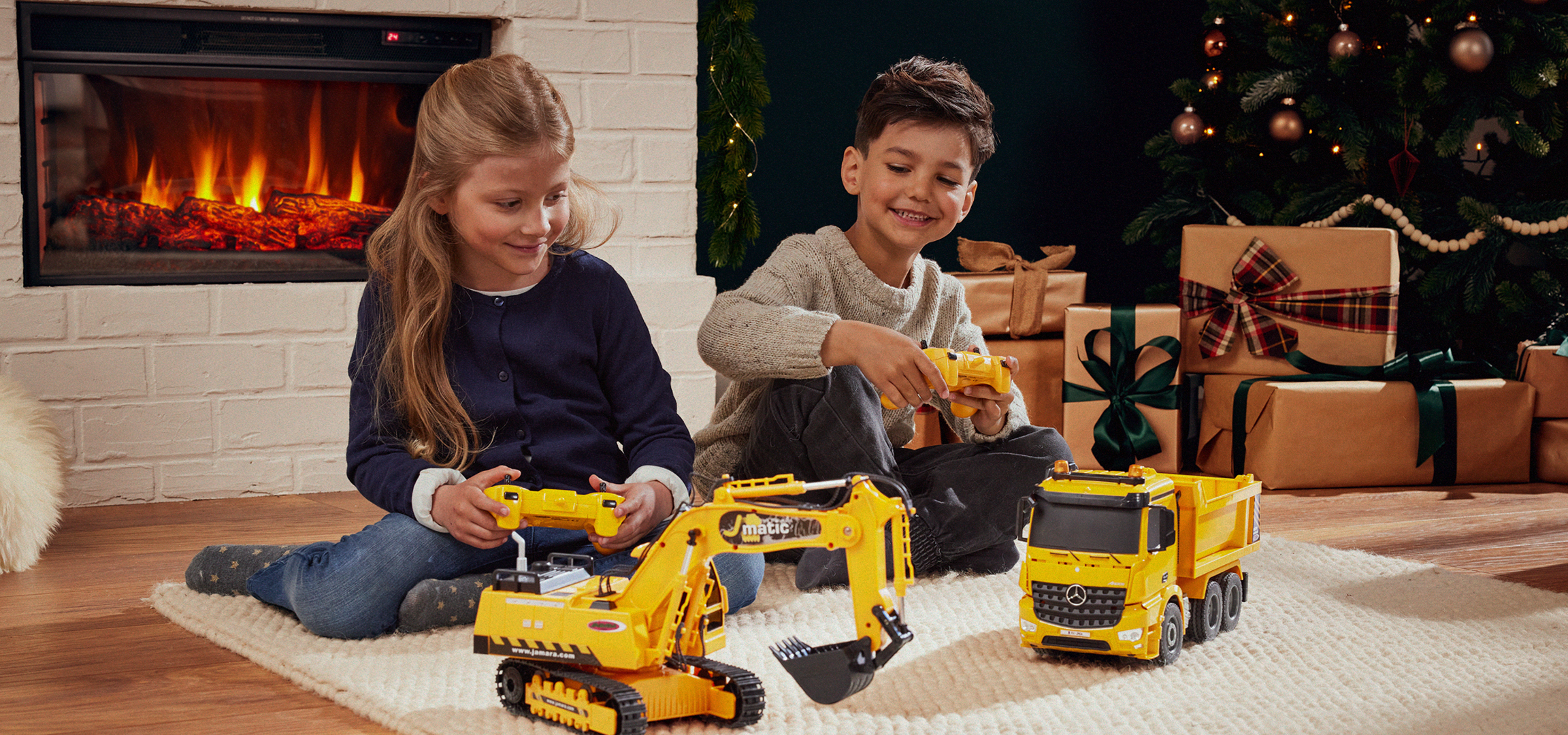 Két gyermek játszik sárga Jamara építőipari járművekkel egy karácsonyfa és kandalló előtt.