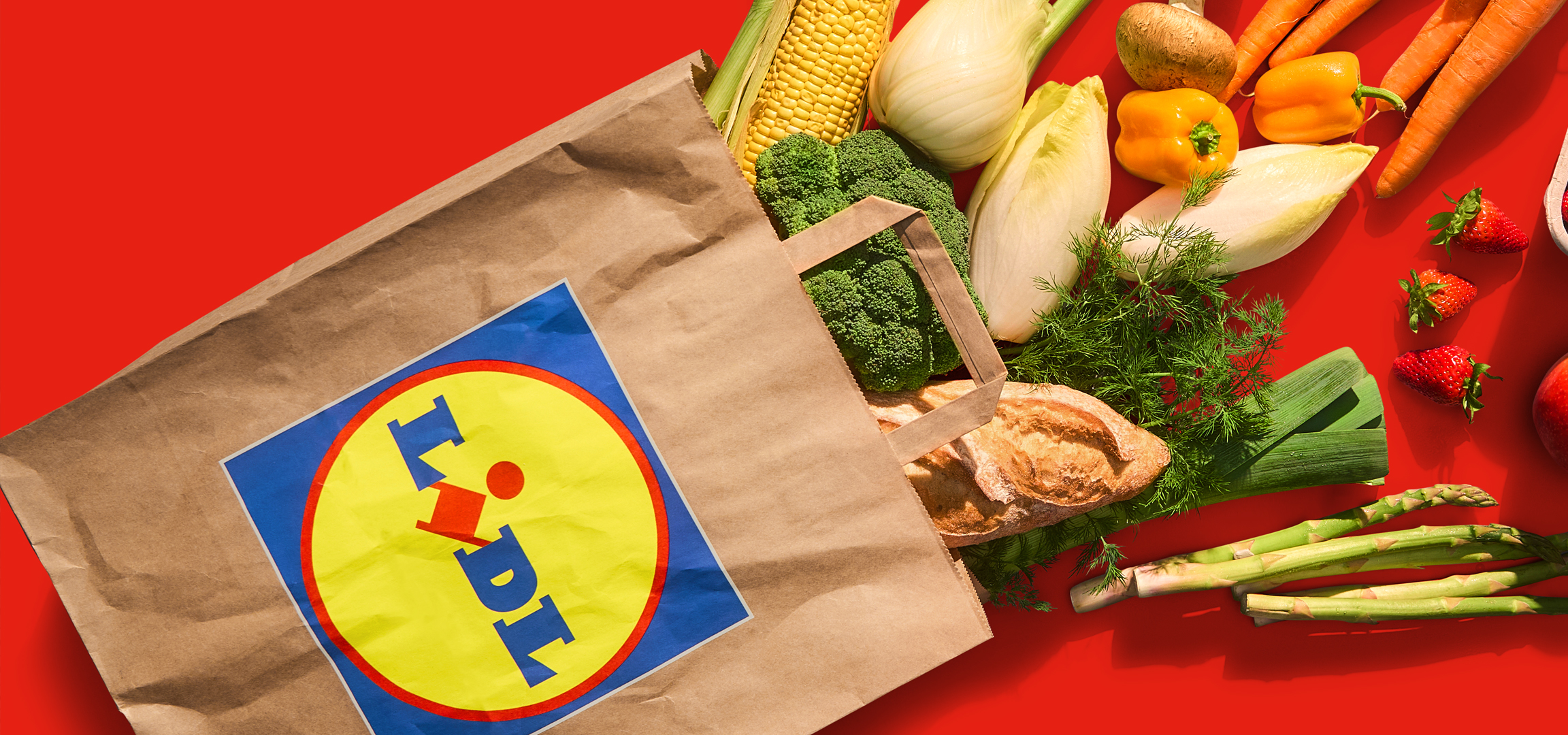 Lidl bevásárlótáska friss zöldségekkel és kenyérrel, piros háttéren.
