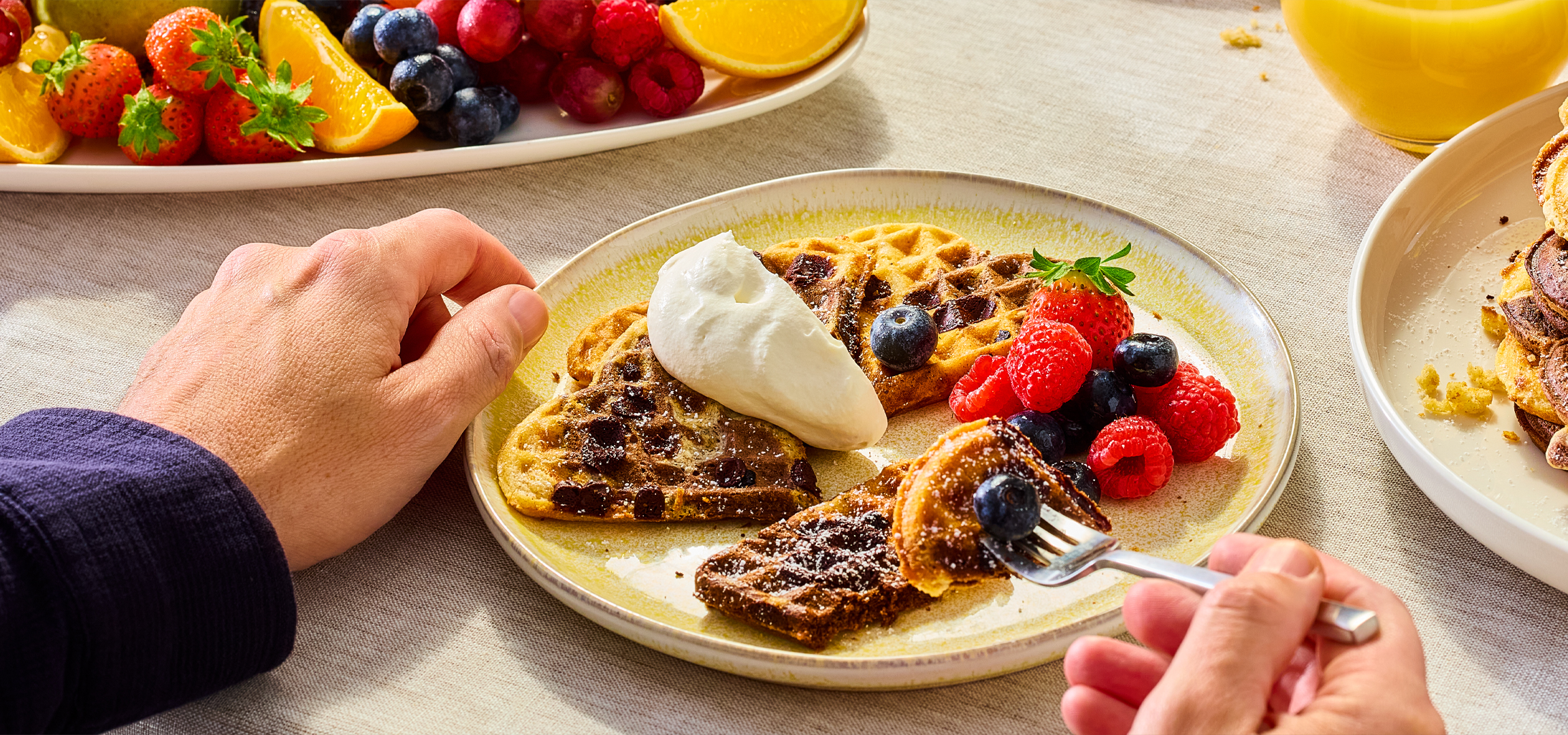 Kéz, amely gofrit eszik tejszínhabbal és bogyós gyümölcsökkel, gyümölcstál és gofrihalom mellett.