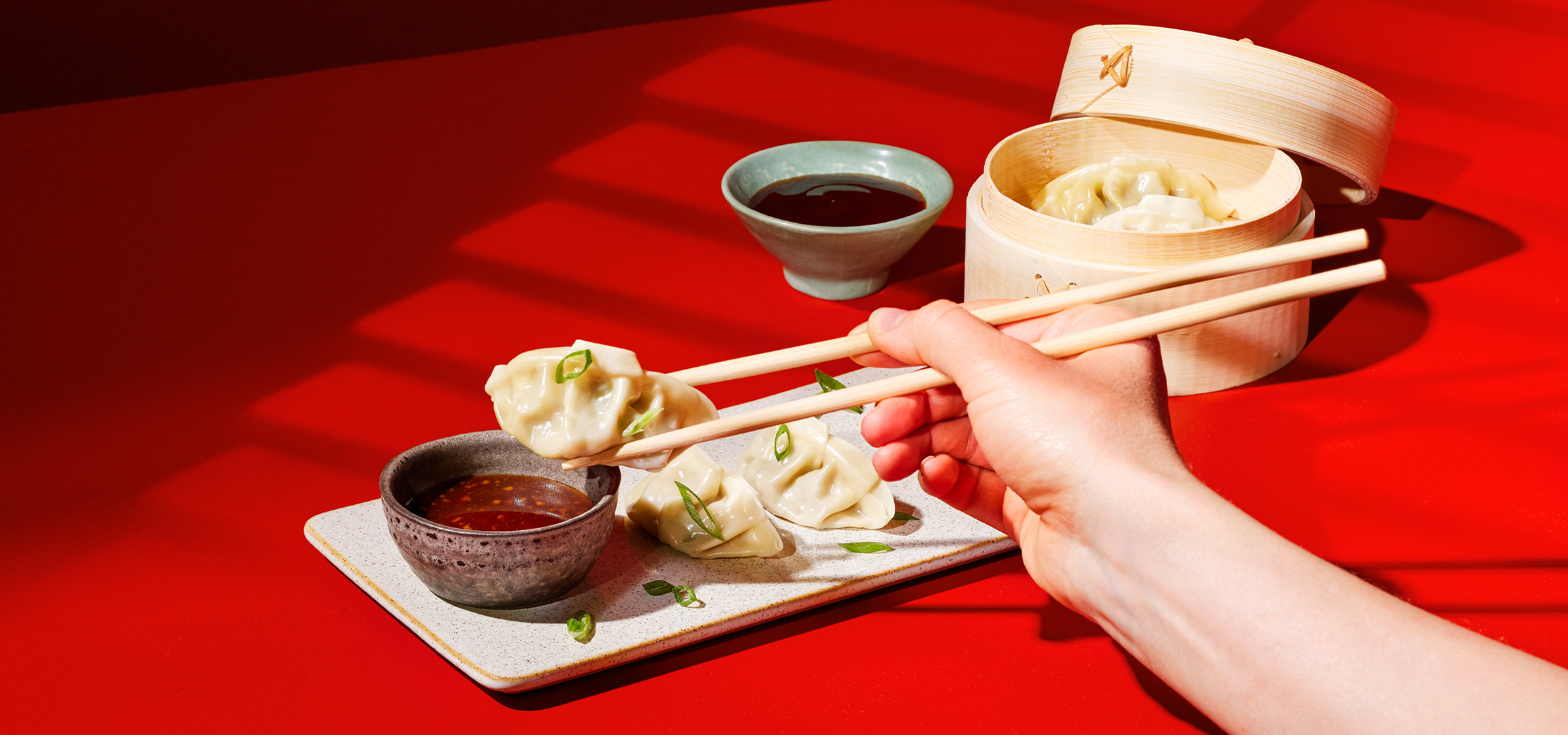 Gombócok mártogatóssal és pálcikákkal, bambusz gőzölőben, piros háttérrel.