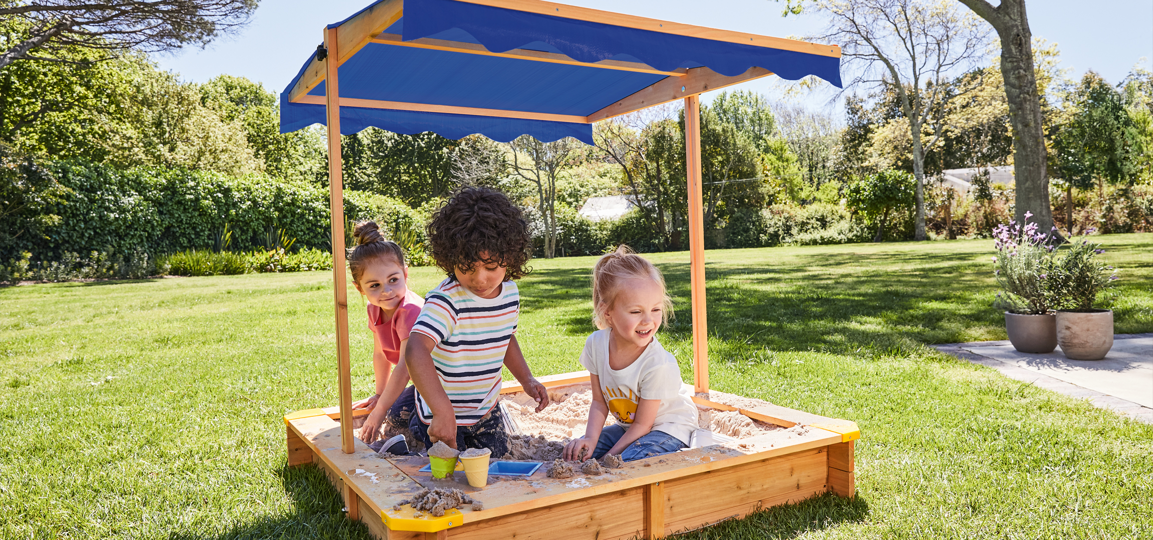 Gyerekek játszanak egy homokozóban, amelynek kék napellenzője van, egy zöld füves kertben.
