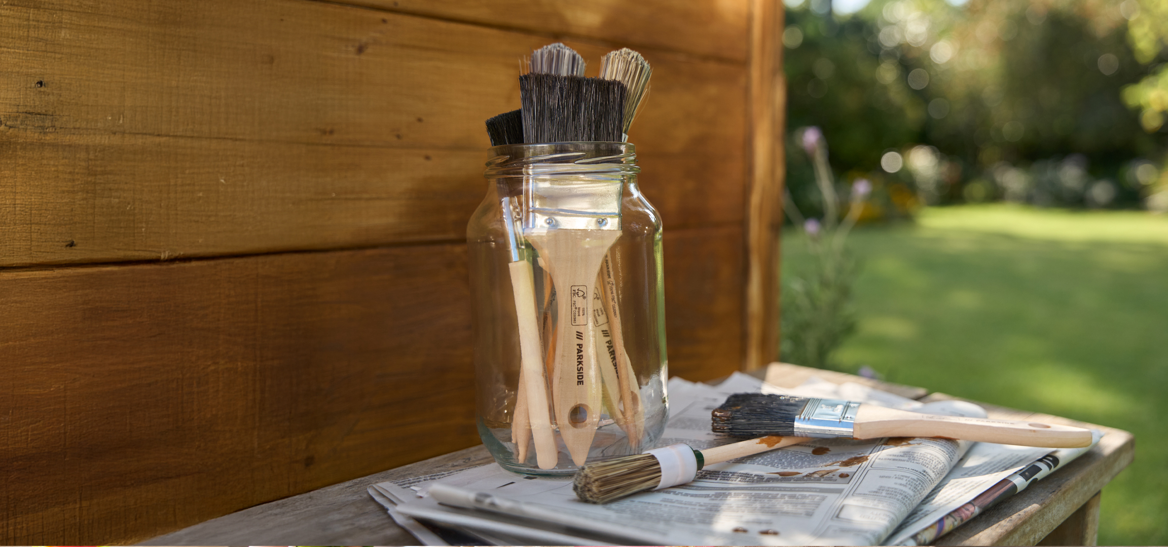 Festőecsetek egy üvegben, újságpapíron, fa háttérrel és elmosódott kerti környezettel.