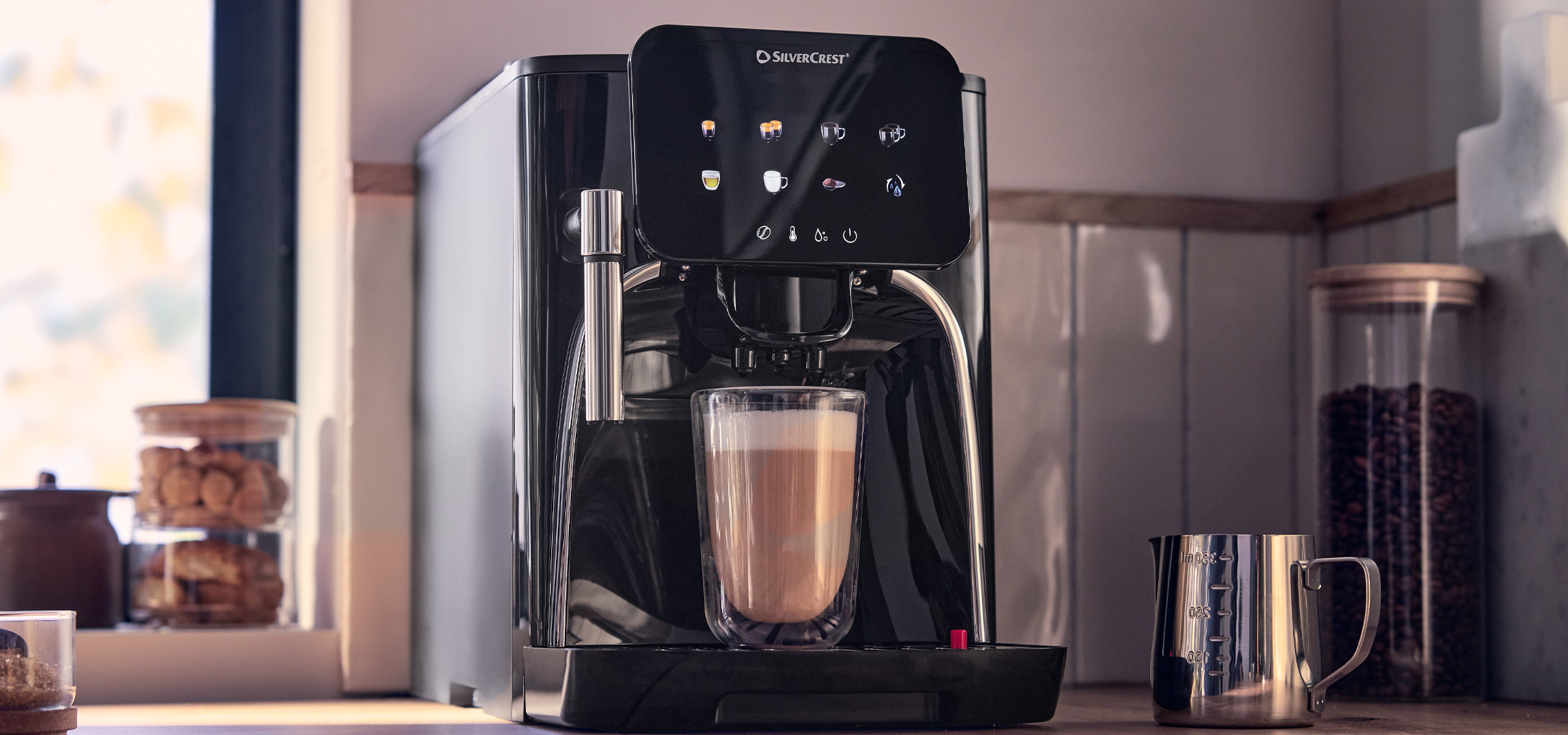 Kávéfőző tejhabos kávéval egy konyhaablak előtt, süteményekkel és kávébabokkal.