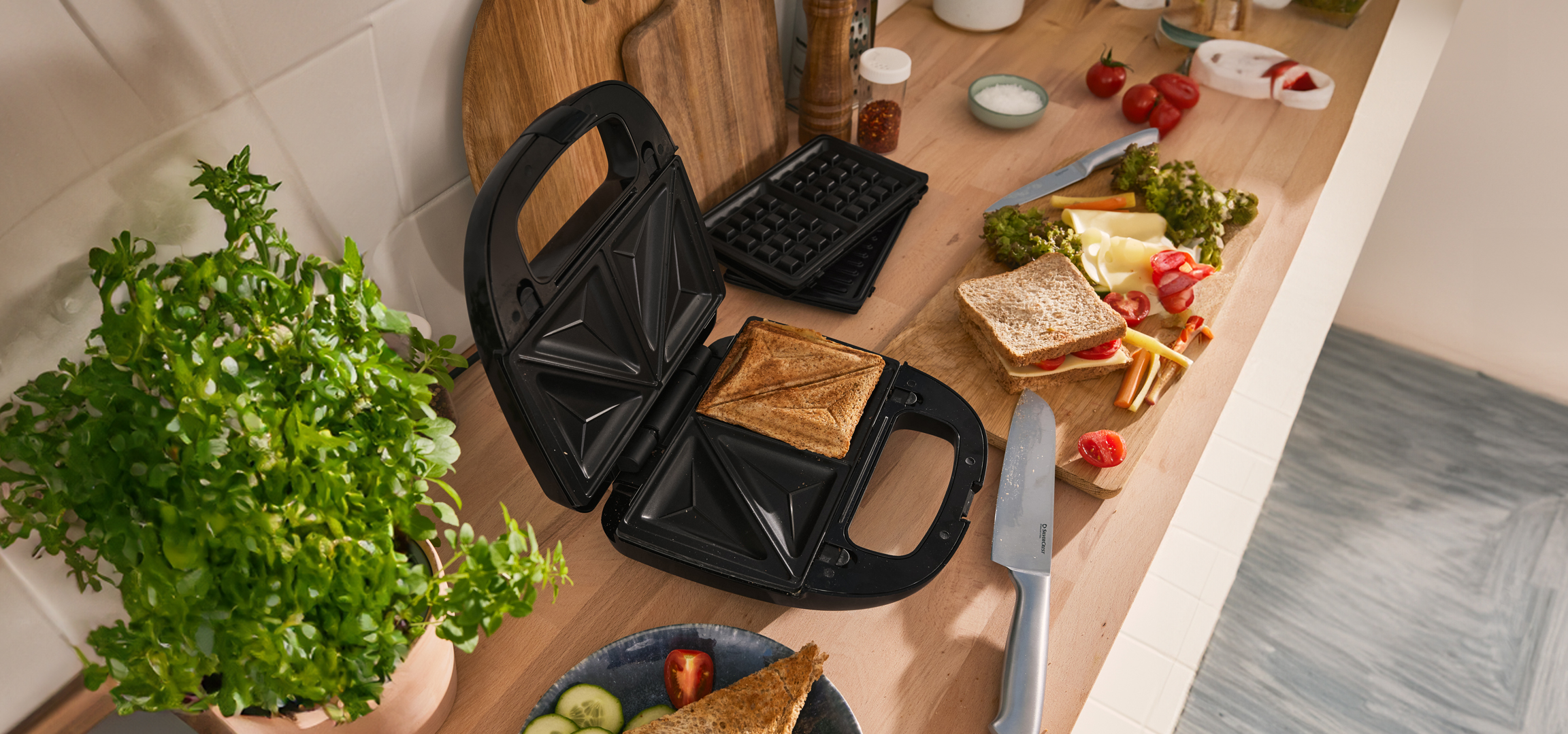 Szendvicssütő nyitva, benne pirított szendvics, mellette zöldségek és sajt egy vágódeszkán.