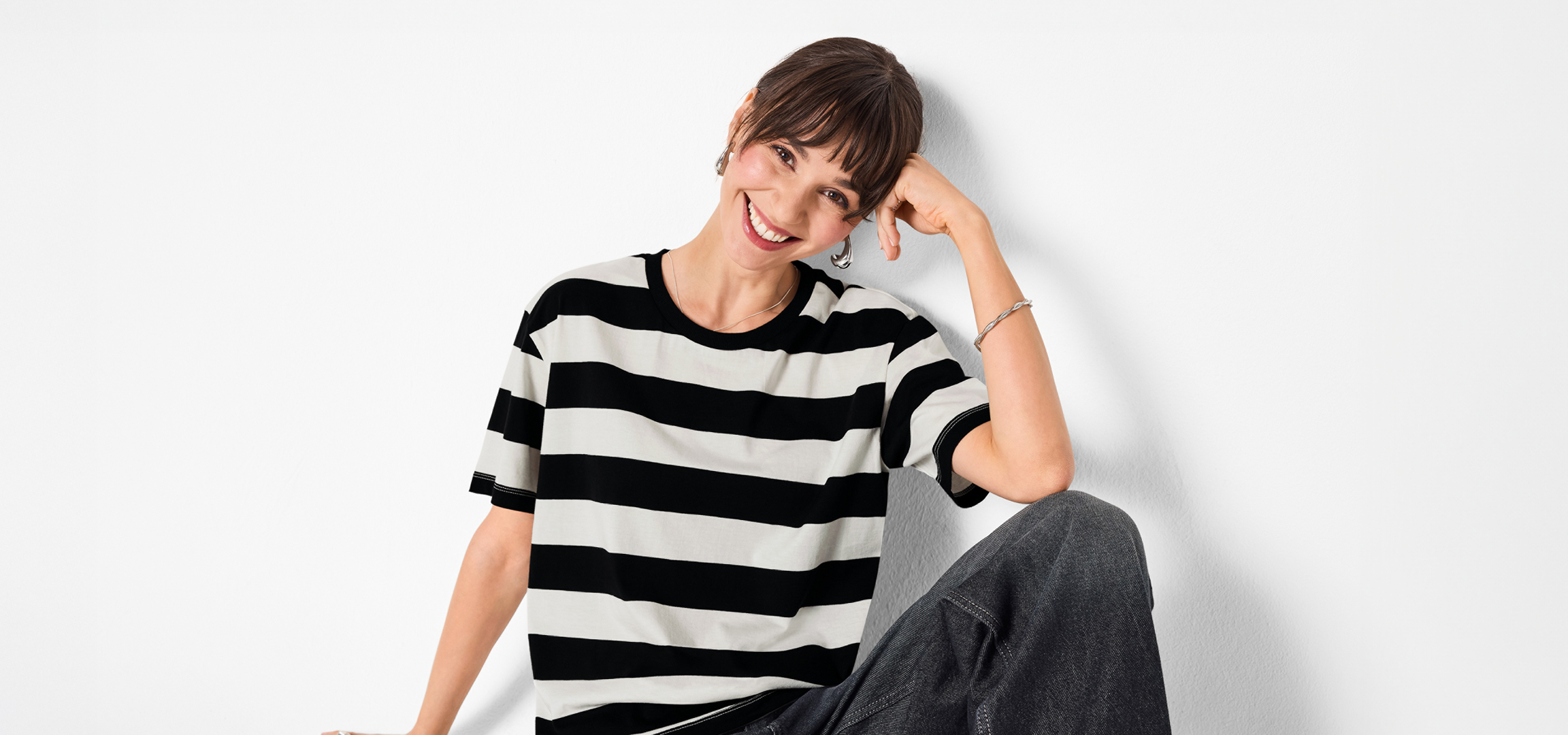 Nő fekete-fehér csíkos pólóban és farmerben, mosolyogva támaszkodik a falnak.