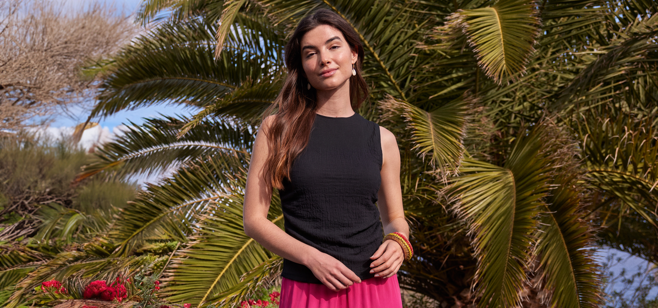Nő fekete ujjatlan felsőben és rózsaszín szoknyában pálmafák előtt.