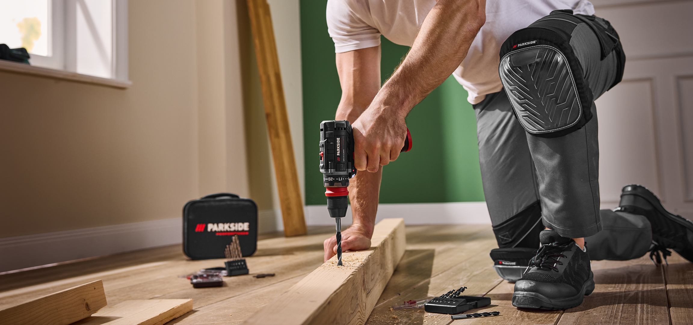 Férfi fúrógéppel fúr egy fadarabot, mellette Parkside szerszámos táska és fúrófejek.