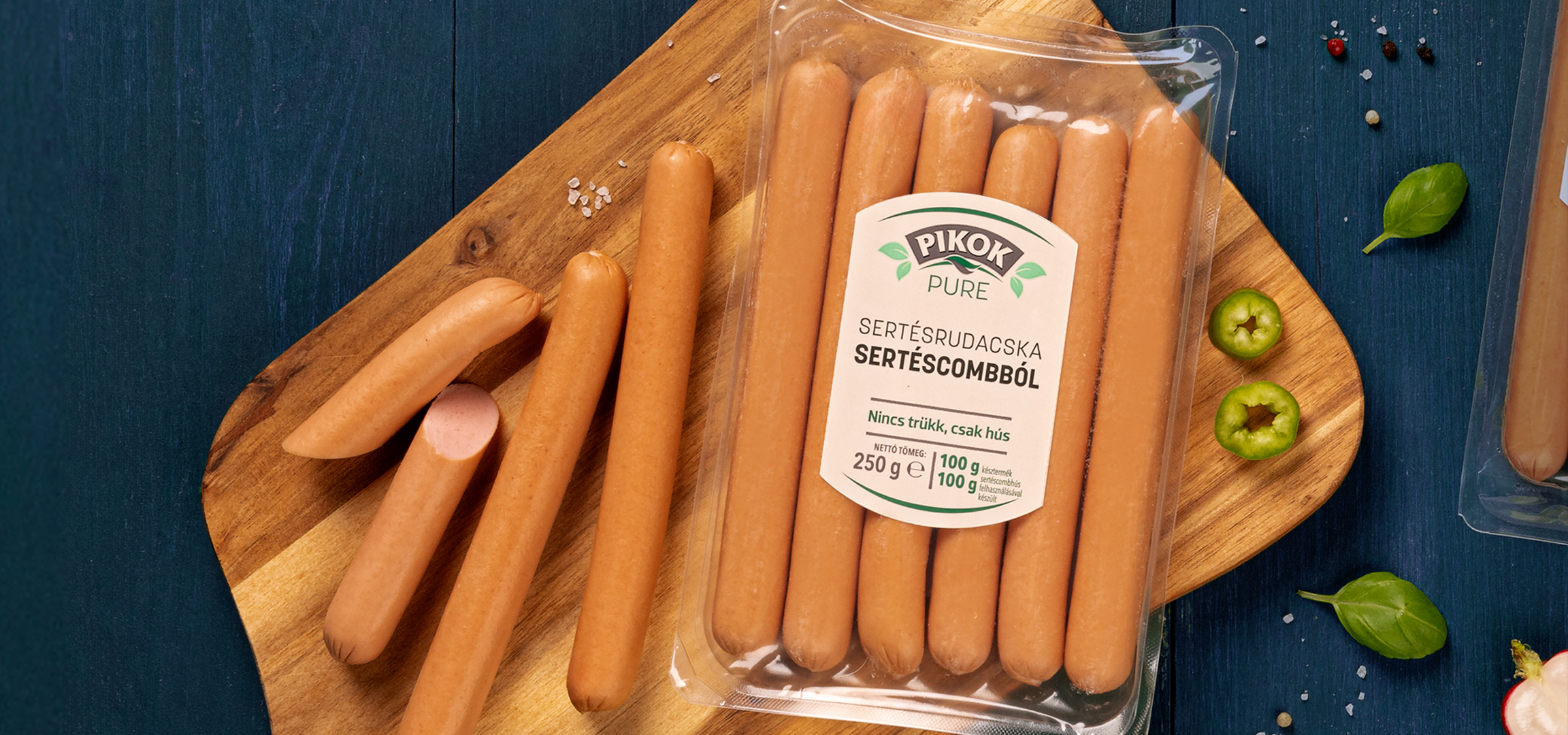 Pikok Pure sertésrudacskák és sertéscombból készült virslik, fűszerekkel és zöldségekkel egy sötétkék fa asztalon.