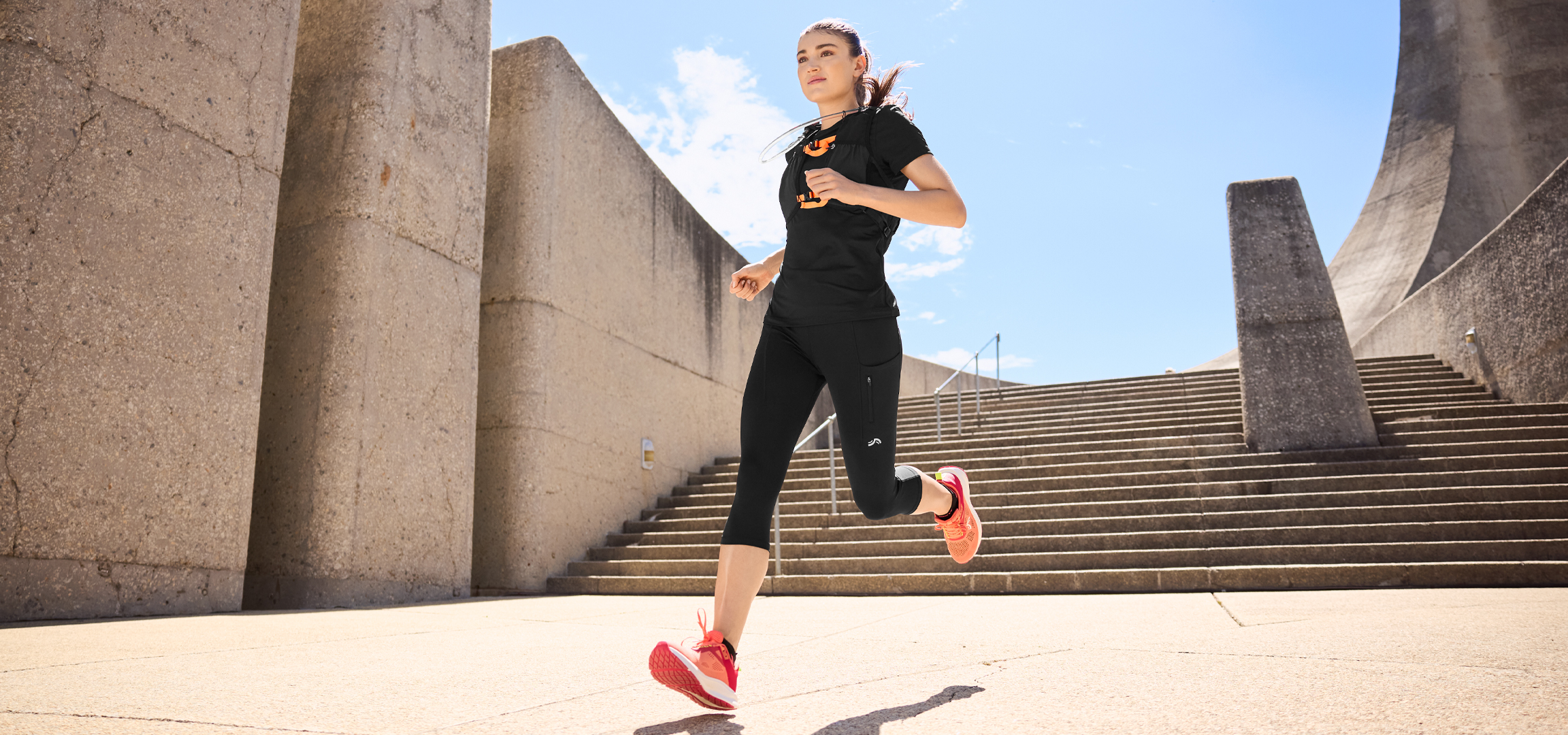Nő futóruhában, fekete pólóban és nadrágban, narancssárga futócipőben.