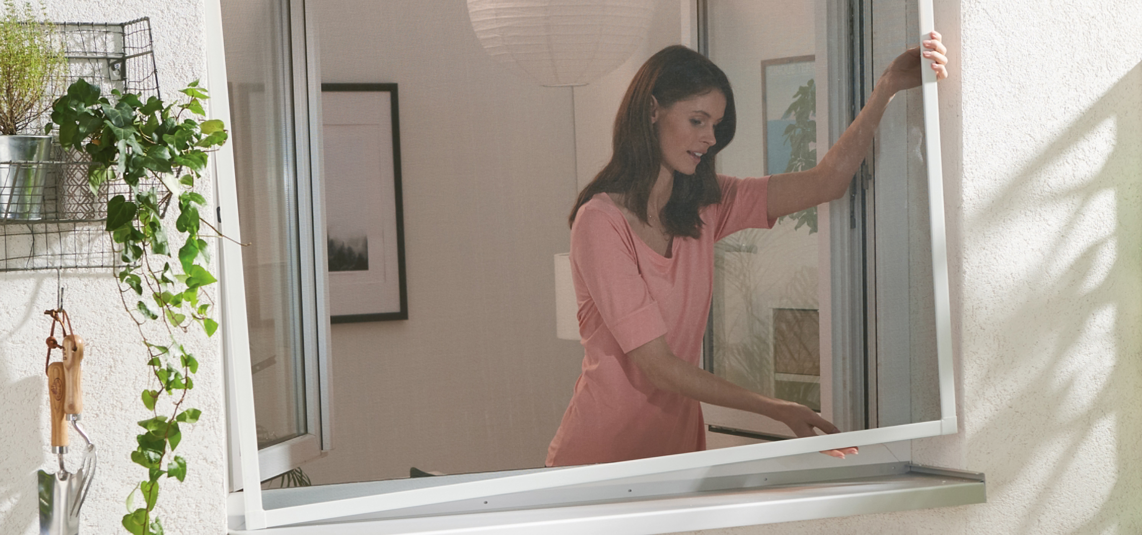 Nő szerel fel egy szúnyoghálót egy ablakra, mellette növények és kerti szerszámok.