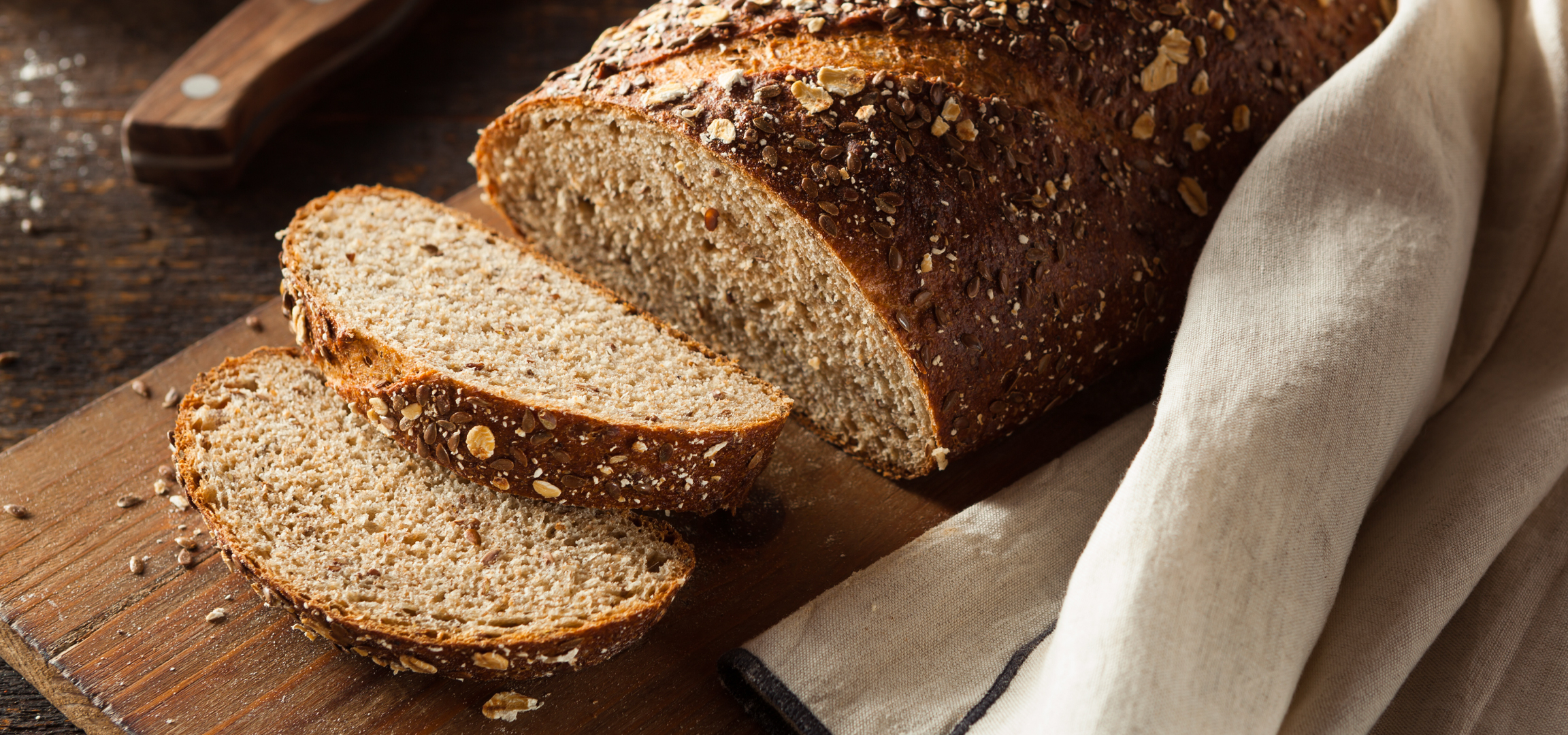 Szeletelt, magvas kenyér egy vágódeszkán, mellette egy kés és egy vászonkendő.