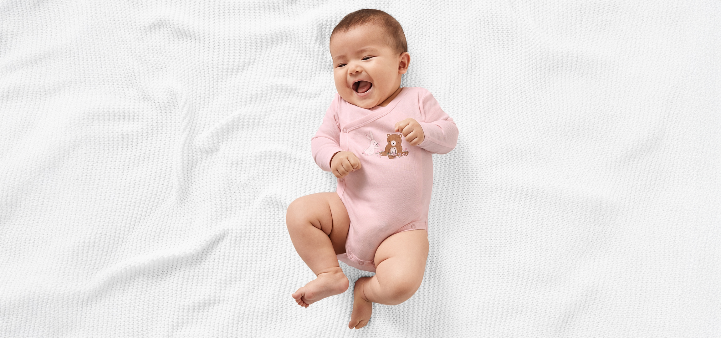 Rózsaszín bodyt viselő baba fekszik egy fehér takarón, tágra nyílt szájjal.