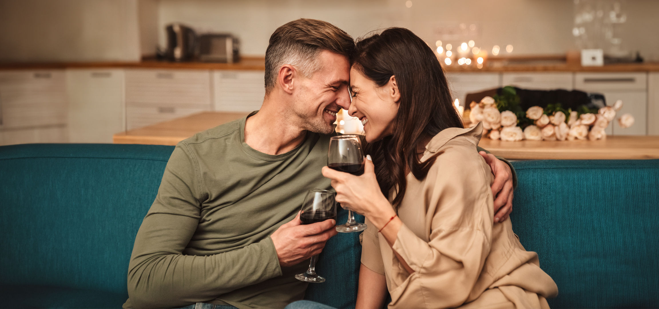 Mosolygó pár vörösborral a kanapén, gyertyákkal és virágokkal a háttérben.
