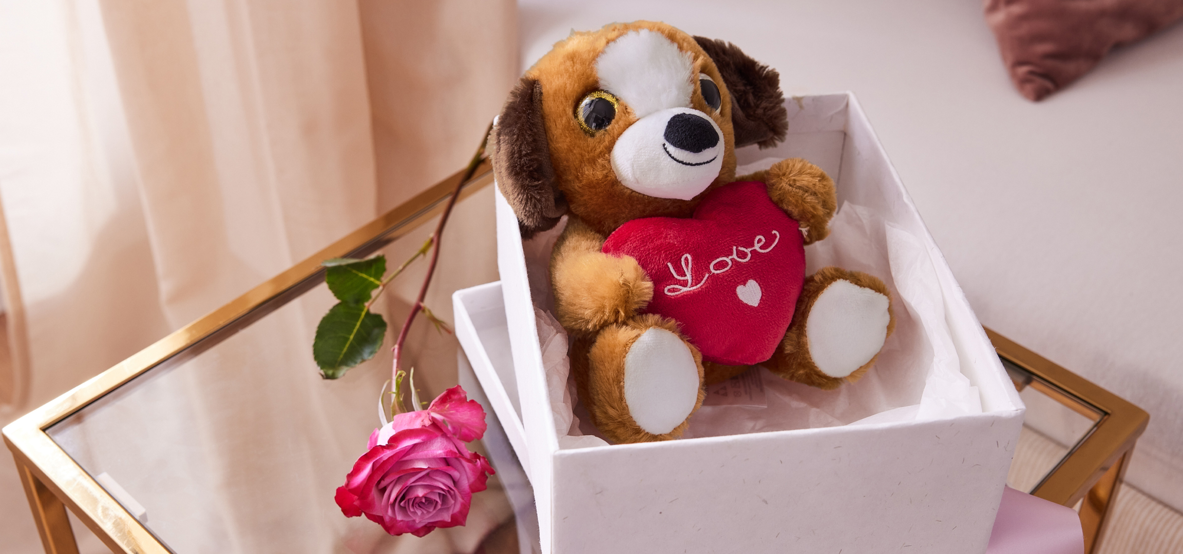 Plüss kutya szívvel és rózsaszín rózsa egy üveg asztalon, világos függönyök előtt.