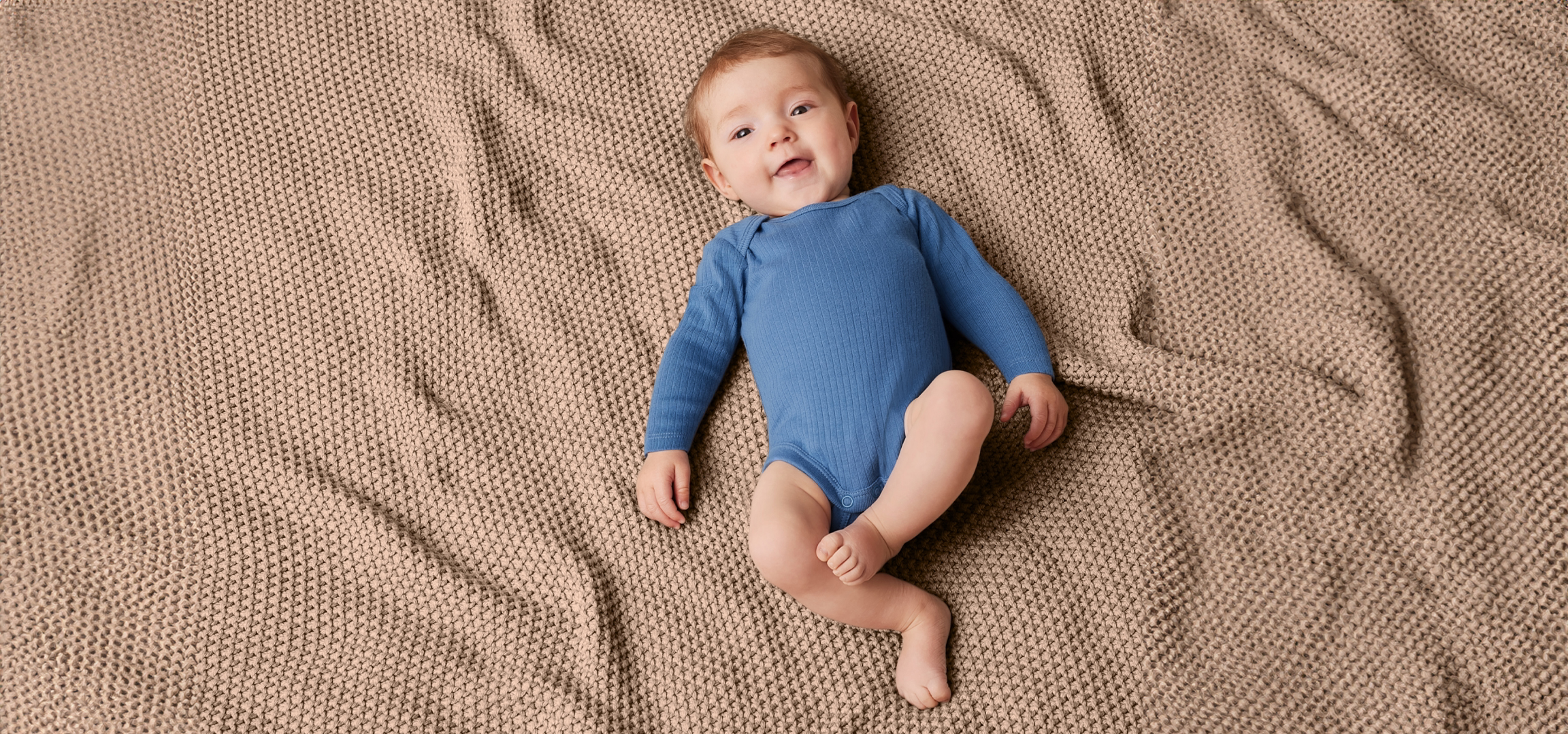 Egy csecsemő fekszik egy bézs színű, kötött takarón, kék bodyban.