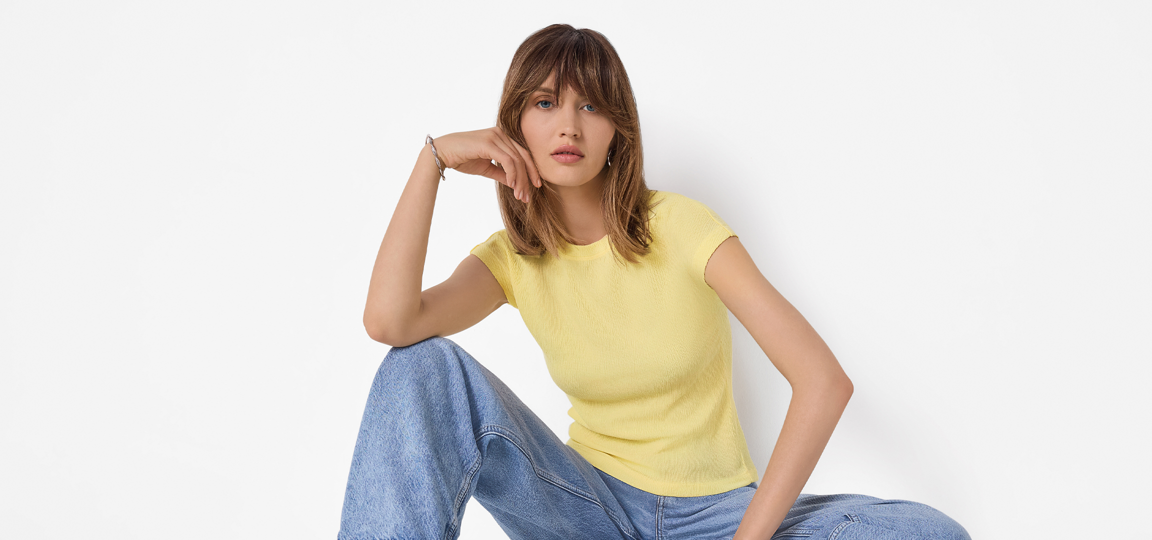 Fiatal nő sárga pólóban és farmerben ül, kezét az arcán pihenteti.