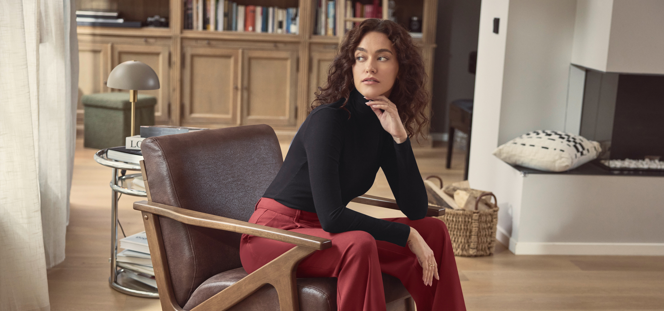 Nő fekete garbóban és piros nadrágban, barna fotelben ülve.