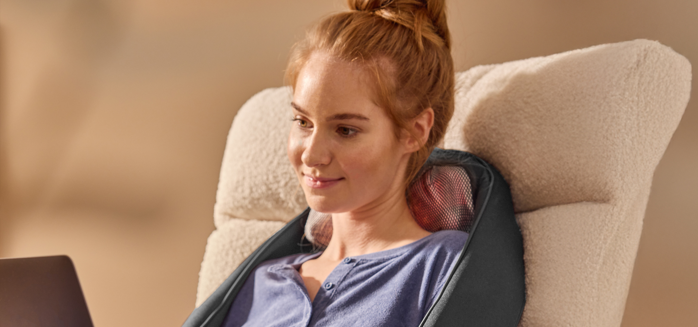 Nő ül egy masszázsüléssel, laptopot használva egy kényelmes székben.