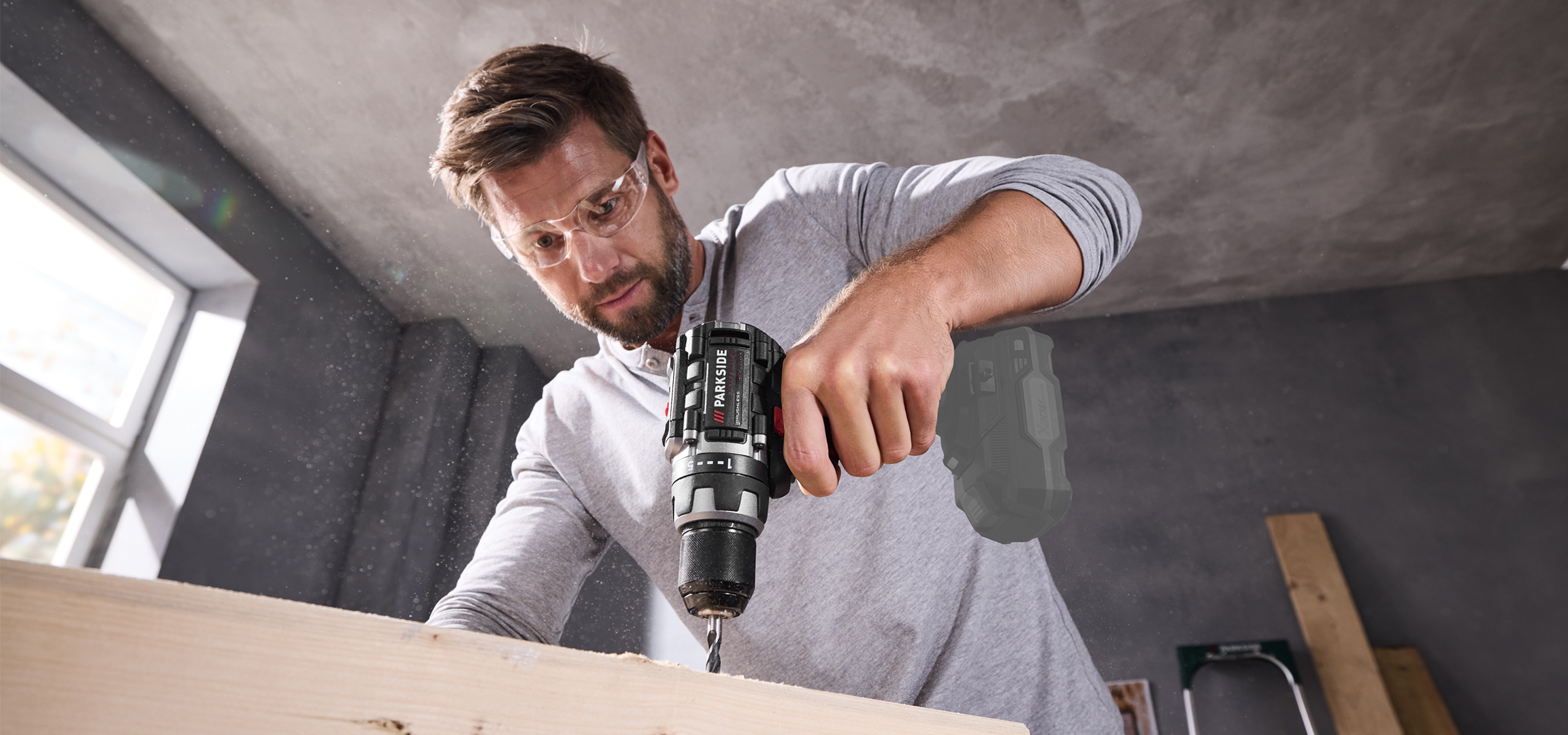 Férfi Parkside akkus fúróval fúr egy fadarabot, védőszemüvegben, műhelyben.