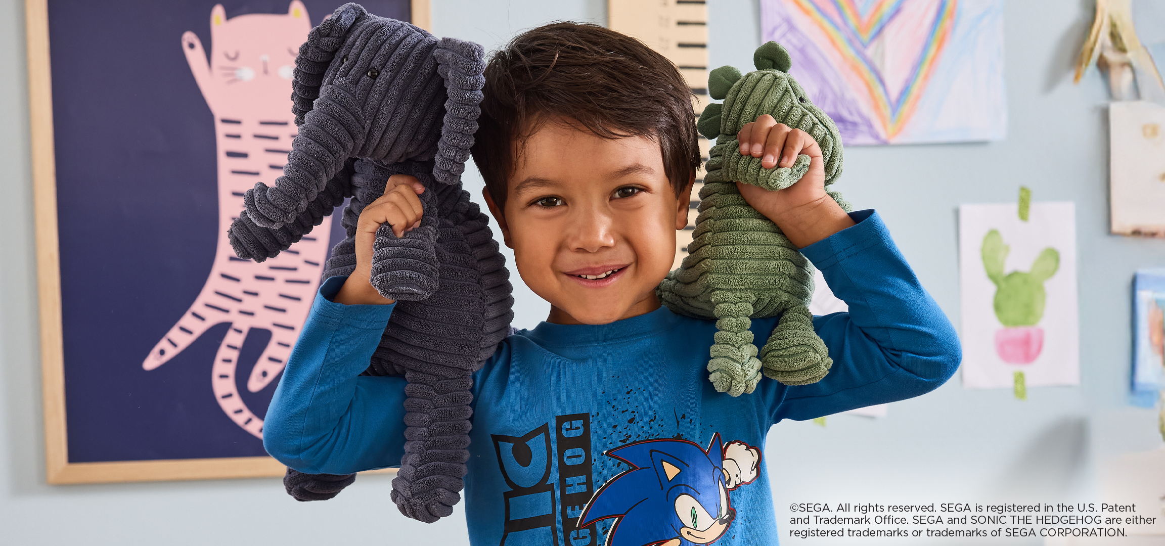 Egy fiú két plüssállatot tart, egy szürke elefántot és egy zöld dinoszauruszt.