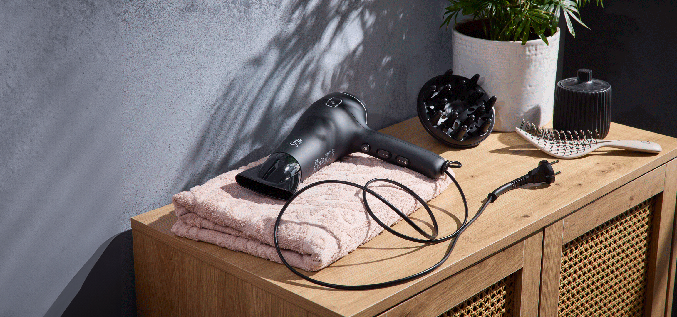 Fekete hajszárító, diffúzor, hajkefe és törölköző egy fa komódon.