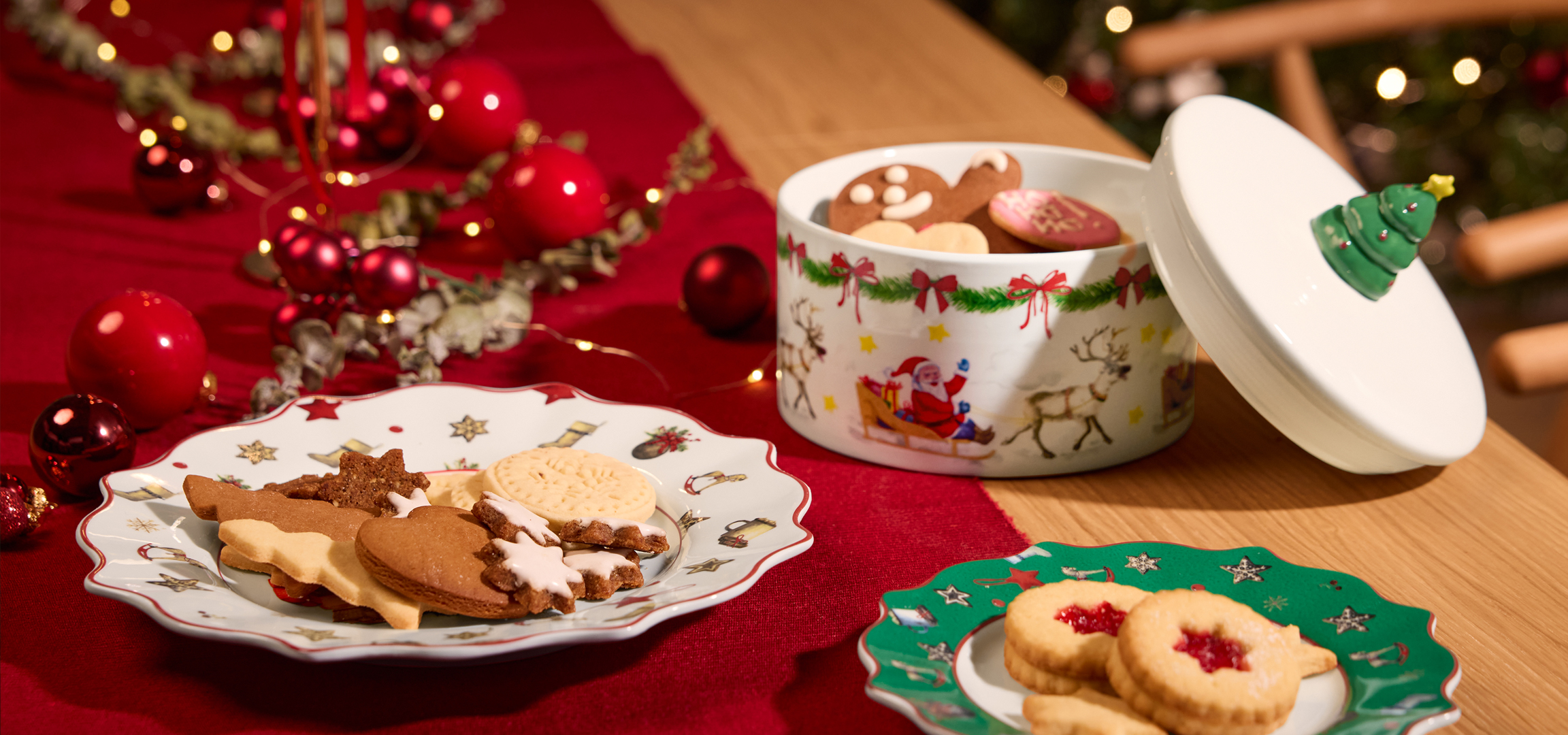 Karácsonyi kekszes doboz mézeskaláccsal és piros karácsonyi díszekkel egy piros terítőn.