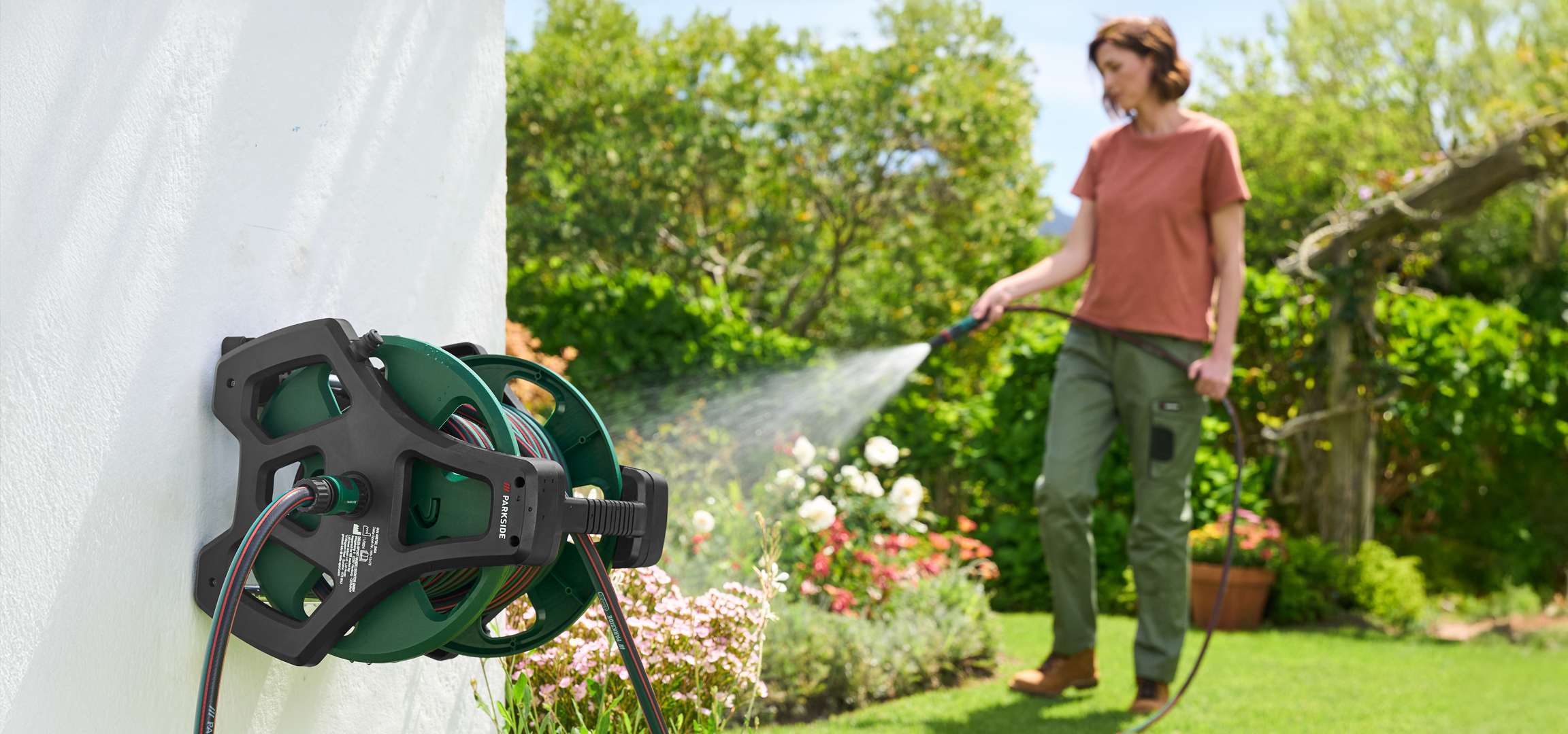 Kerti tömlődob a falra szerelve, egy nő öntözi a kertet